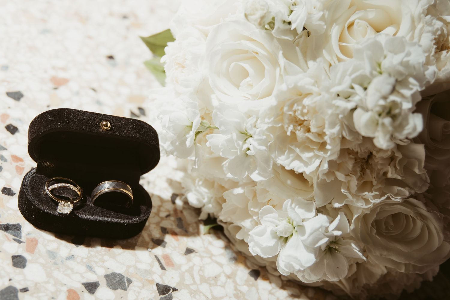 White roses and wedding rings rest on a textured surface creating an elegant bridal detail shot. Wedding Photographer DFW