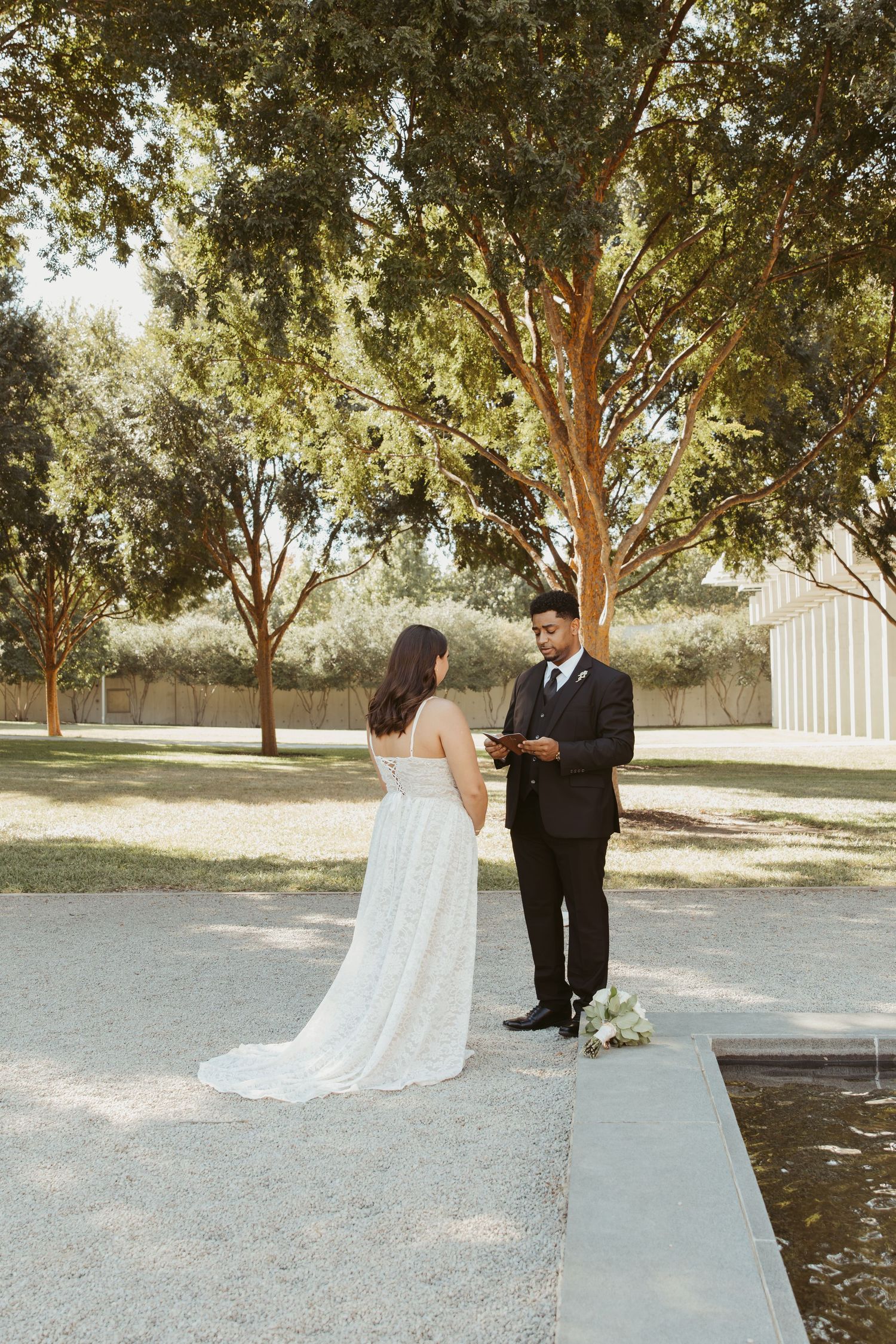 A wedding couple exchanges vows beside a tranquil water feature surrounded by trees in a park setting. Wedding Photographer DFW