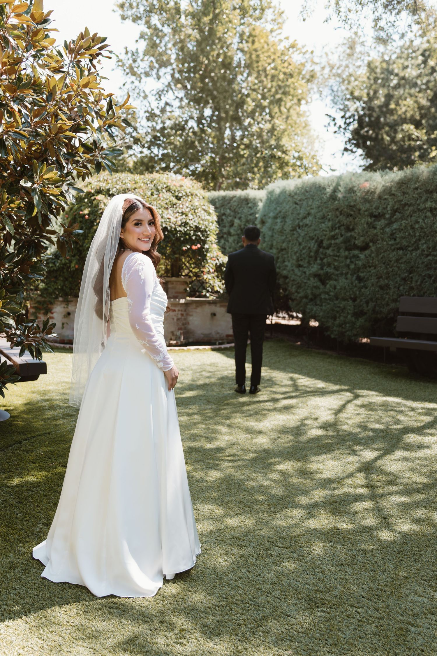 A bride in a white long-sleeved gown looks back at the camera in a garden setting with manicured hedges.