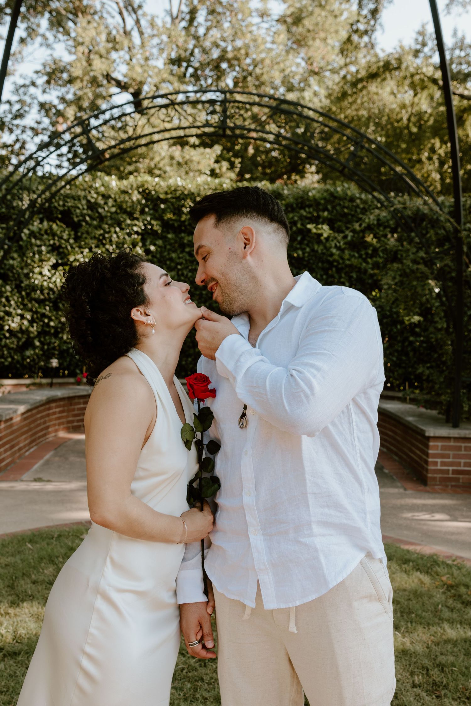 A romantic couple in white outfits shares tender moments together in a sunlit outdoor garden setting.