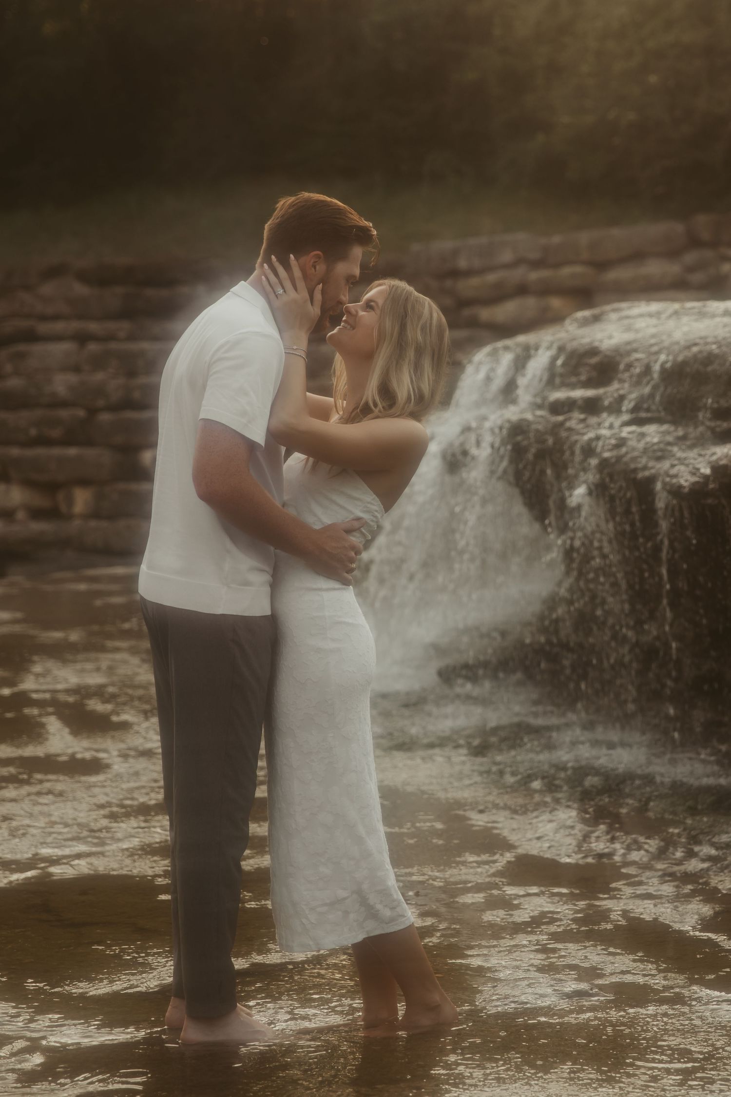 Couple sharing a romantic kiss standing in shallow water in front of a cascading waterfall, wearing white outfits.