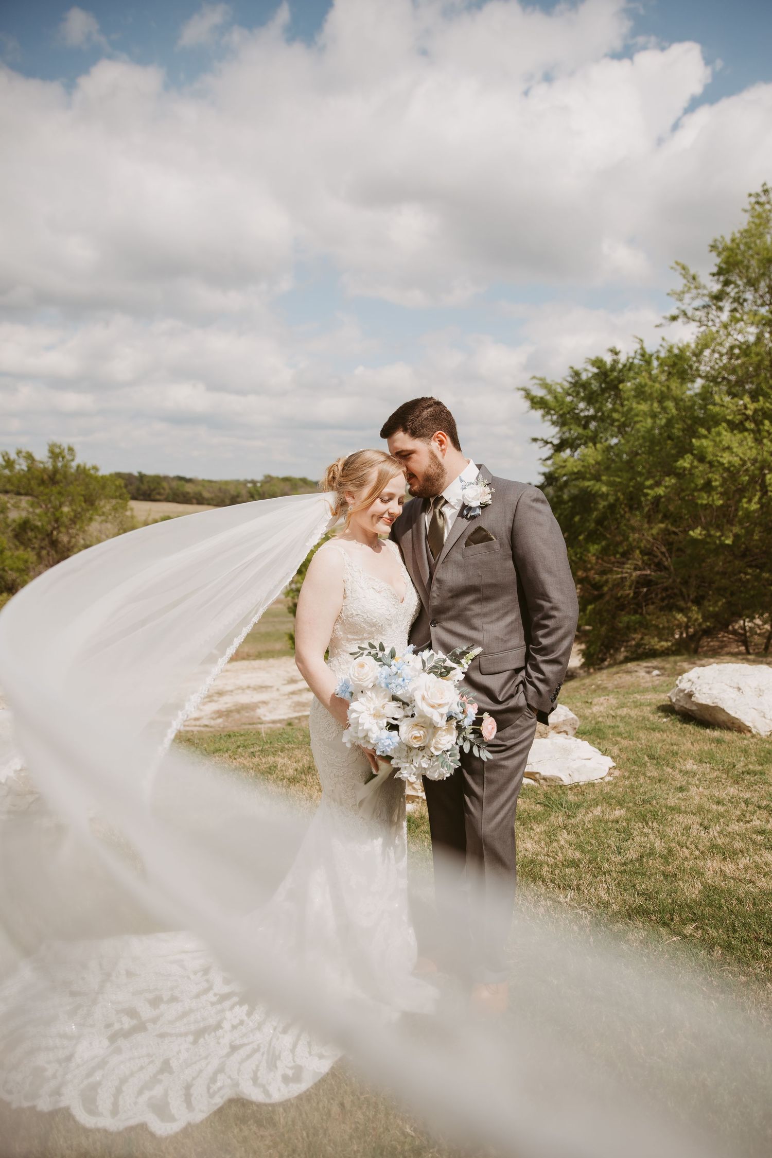 Bride and groom share a romantic moment outdoors as her lace veil billows in the wind on their wedding day.