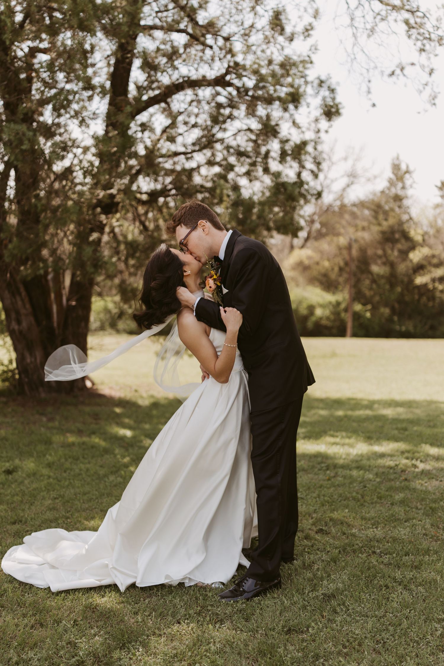 A bride and groom share a romantic kiss outdoors on green grass, surrounded by lush trees on their wedding day.