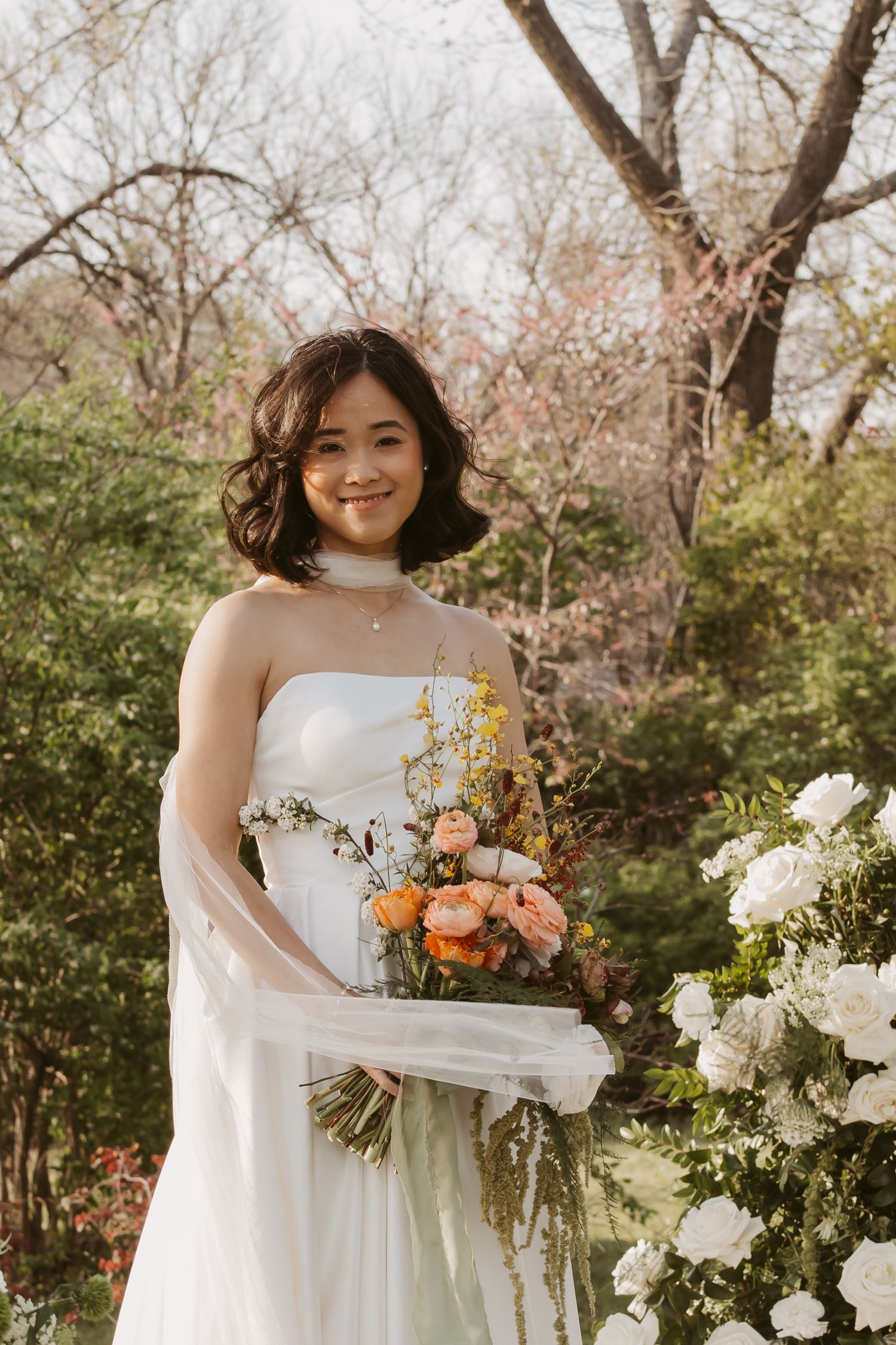Smiling Asian bride in strapless white gown holds colorful orange bouquet beside white floral arrangements outdoors.