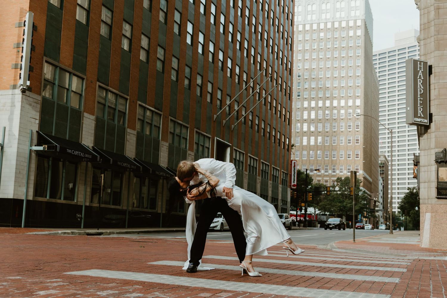Couple in wedding attire share a romantic dip in front of modern skyscrapers on a brick-paved city street.