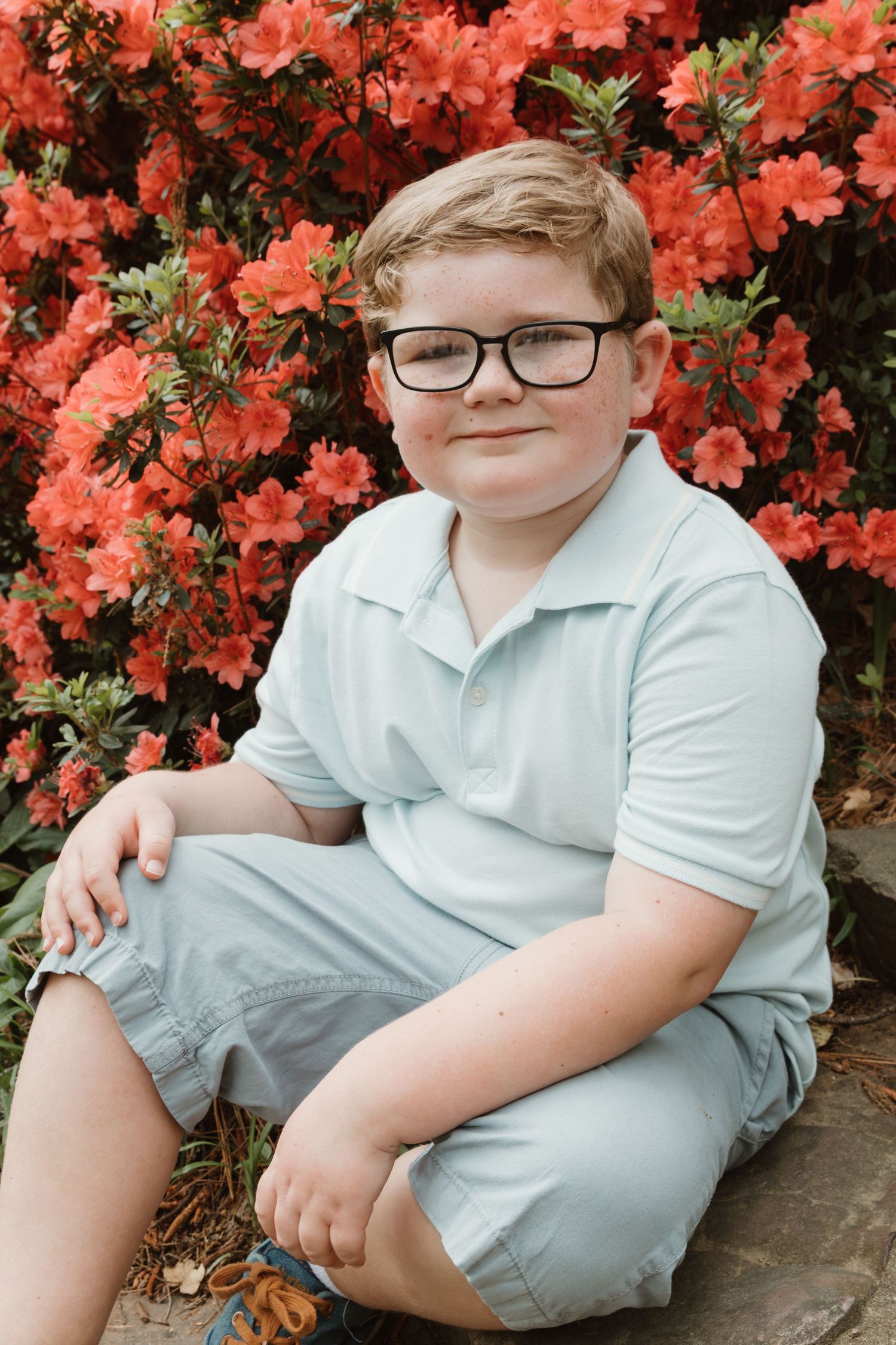 A portrait of a young person in glasses sitting against vibrant coral-colored azalea flowers.