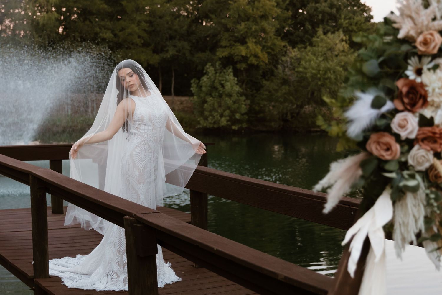 A bride in a flowing white gown poses on a wooden dock surrounded by autumn foliage near a serene lake.