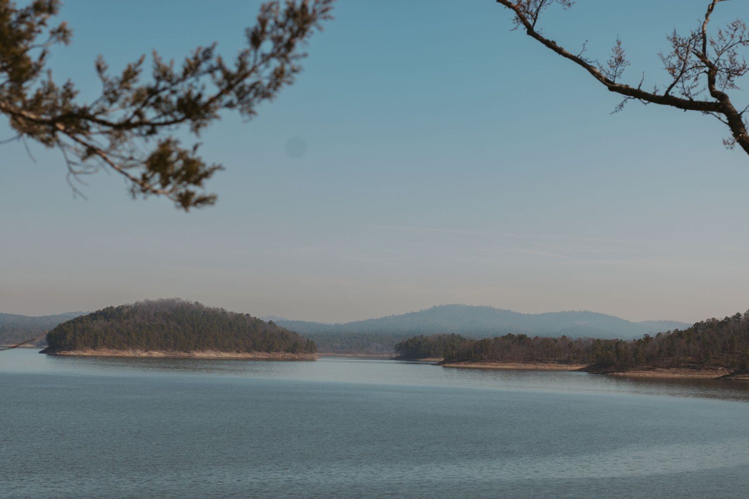 A scenic panoramic view of a mountain lake surrounded by pine trees and distant hills in the background.