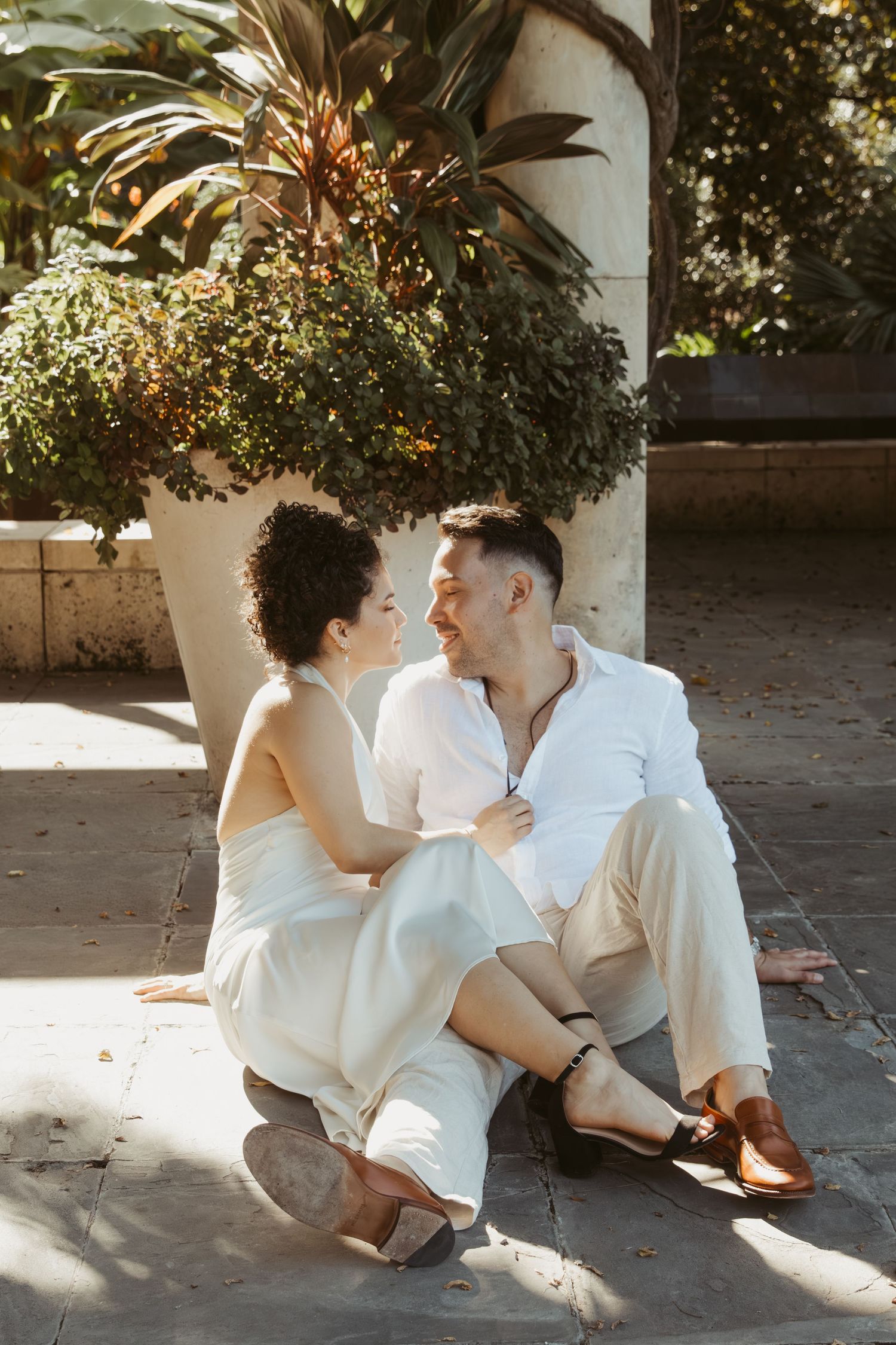 A romantic couple in white outfits sits together on stone steps in dappled sunlight near tropical plants.