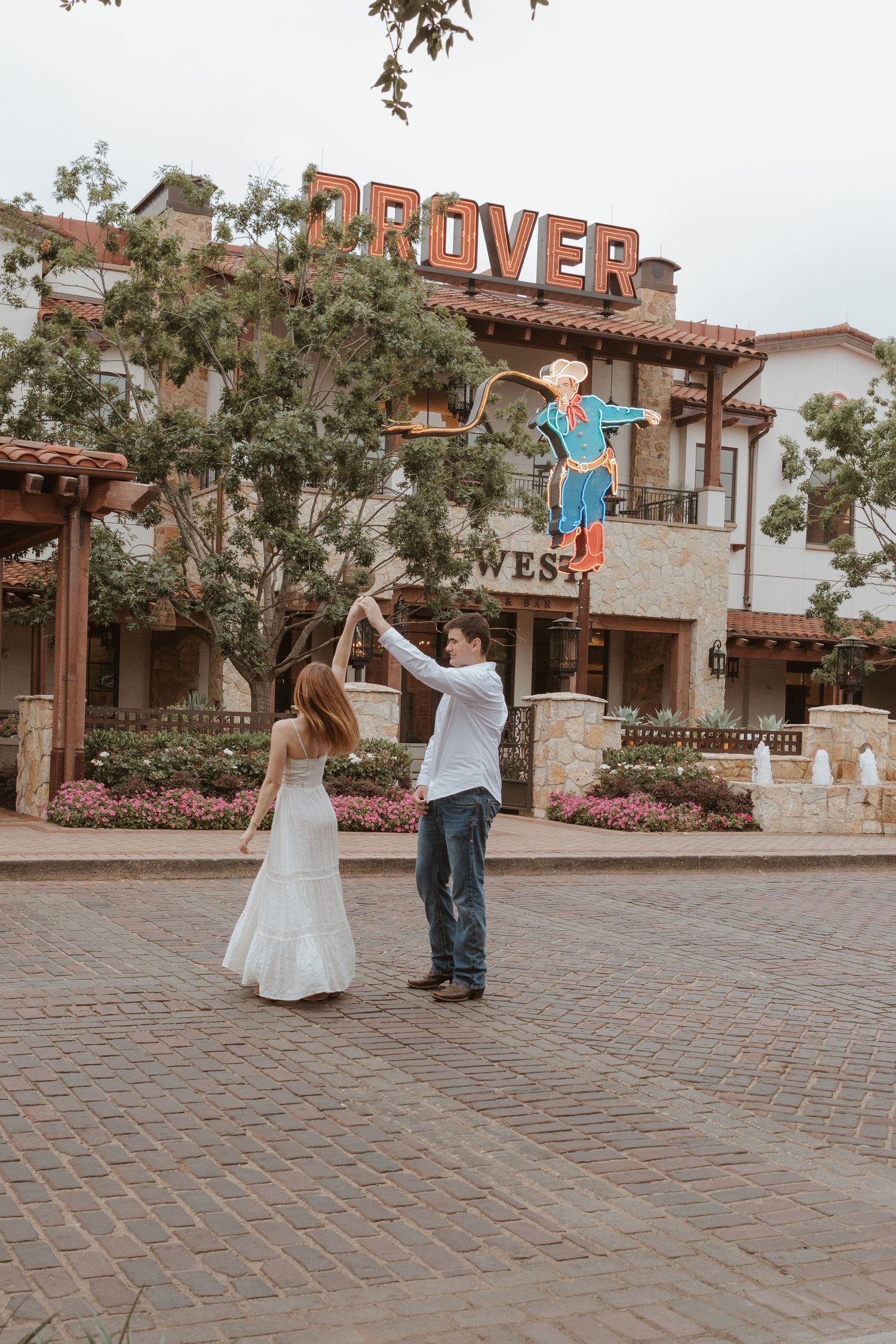 Couple dancing playfully in a rustic courtyard with a vintage storefront and brick paving stones.