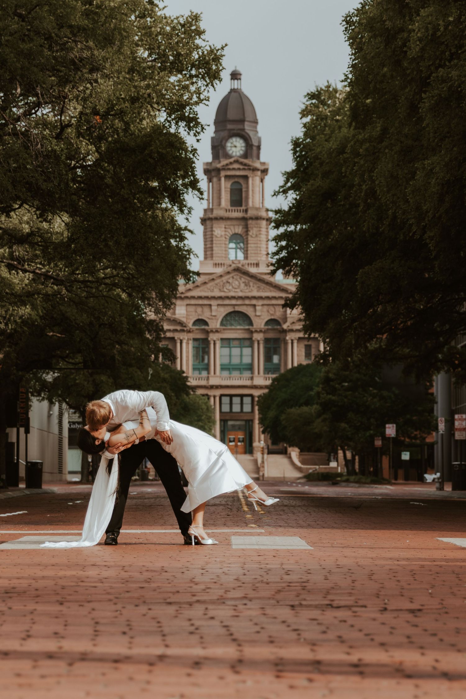 Elegant couple in wedding attire share a romantic dip pose on a brick walkway in front of a historic clock tower building.