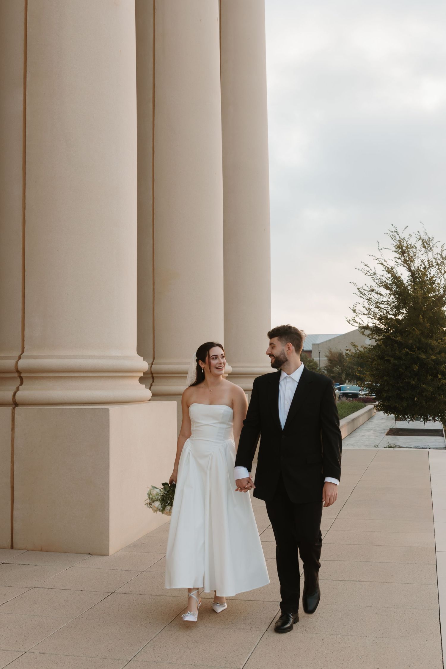 A bride in white and groom in black walk together along a columned walkway during their wedding. Wedding Photographer Fort Worth