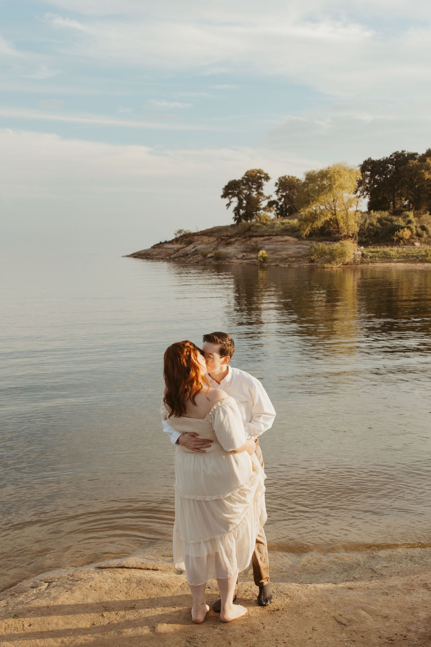 Two people in white clothing embrace on a sandy beach with water and cliffs in the distance.