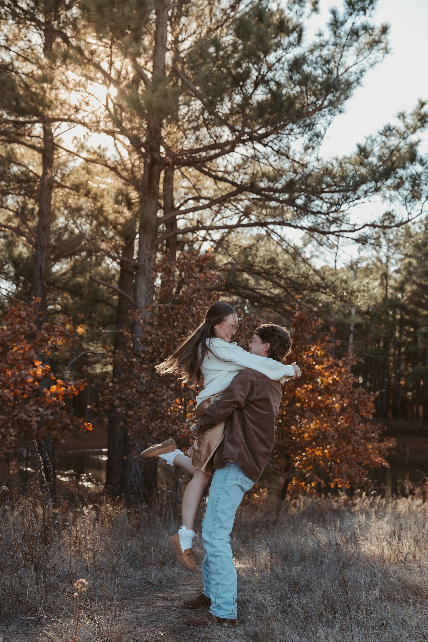 A couple embraces in a romantic dance pose among tall grass and pine trees at sunset. engagement photos DFW