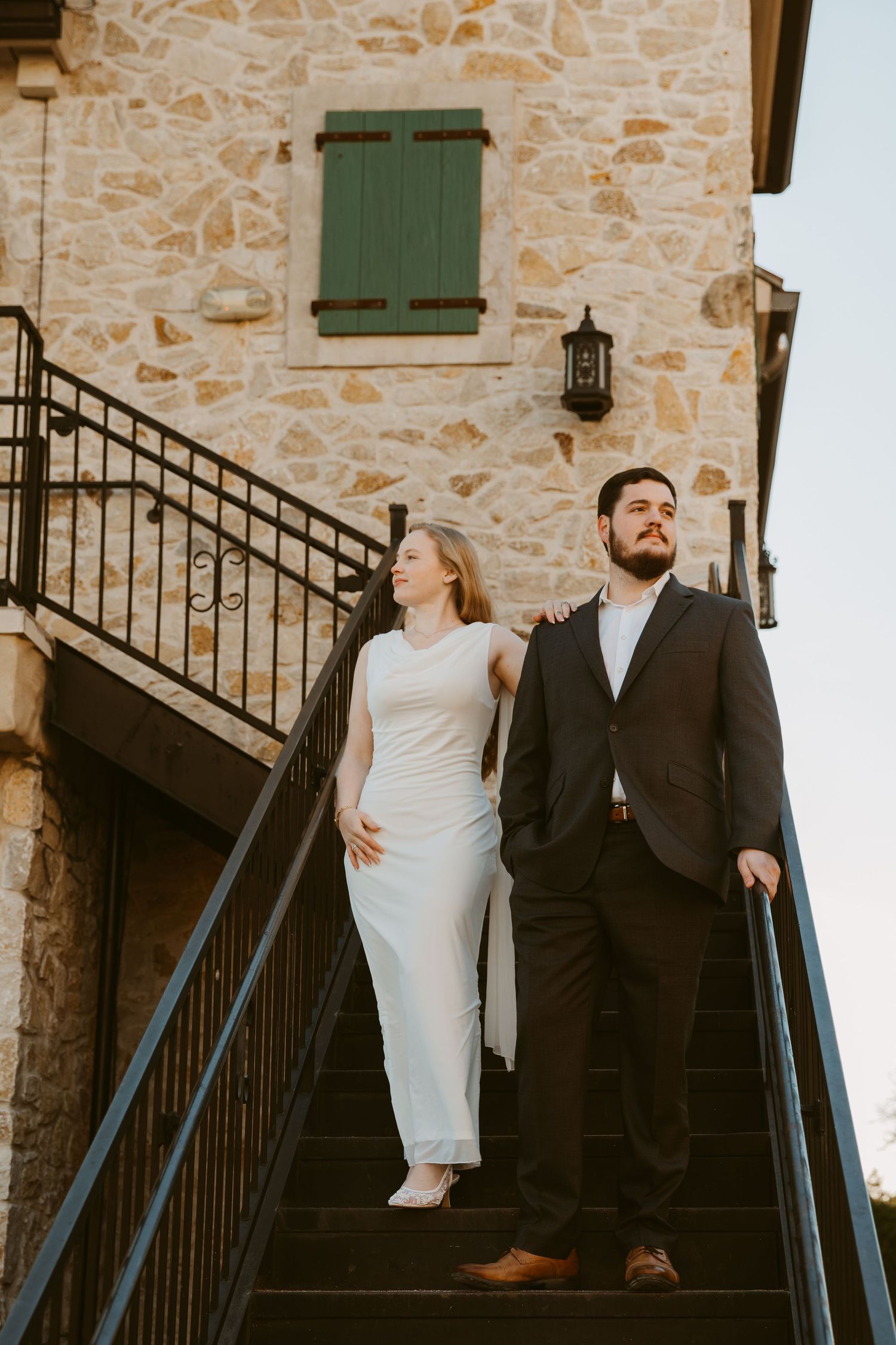 Couple in formal attire poses on stone staircase against rustic building with green shutters.