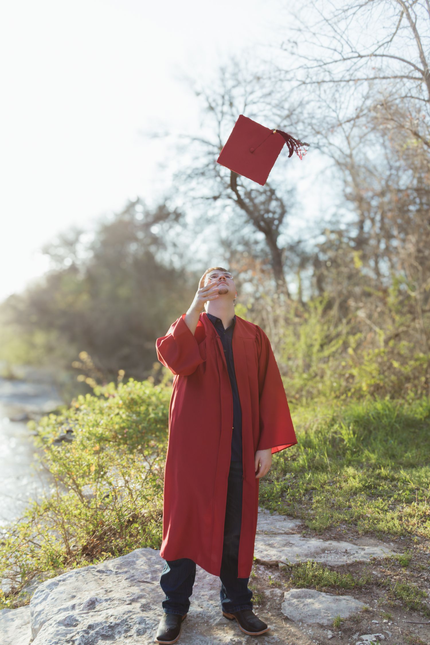 Graduate in red graduation gown celebrates by tossing cap in the air near a river on a sunny day.