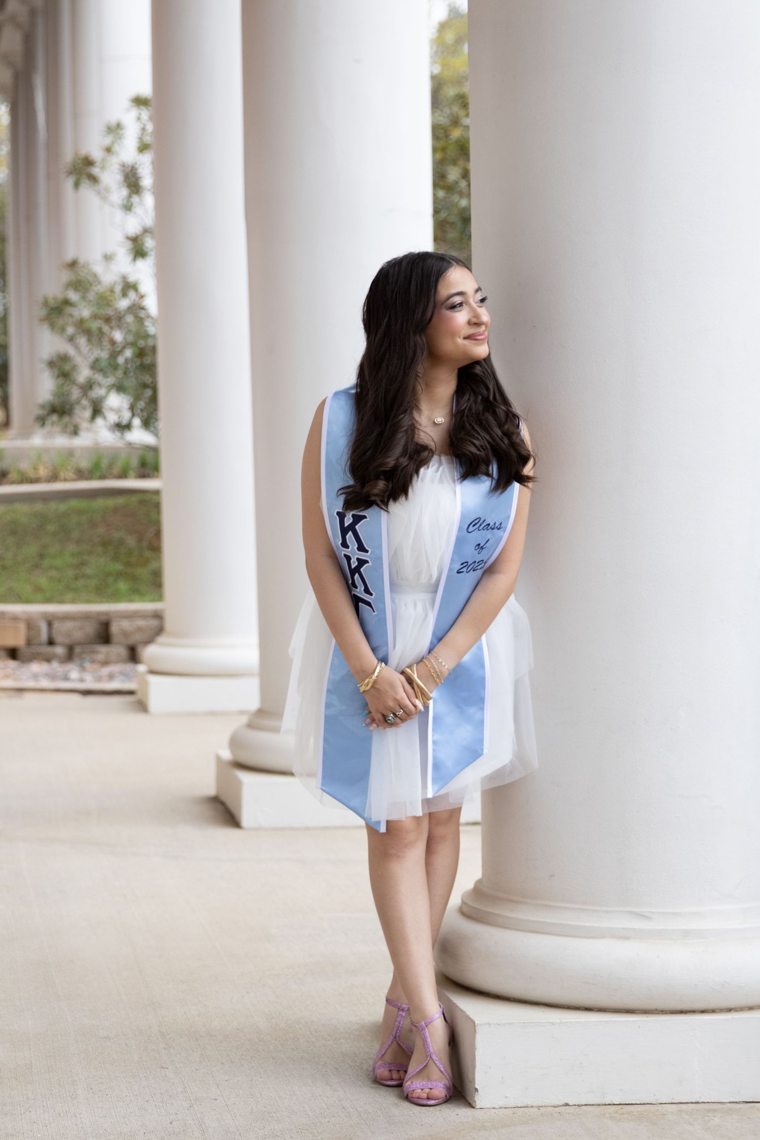 Graduate in a white dress and light blue sash posing elegantly between classical white columns.