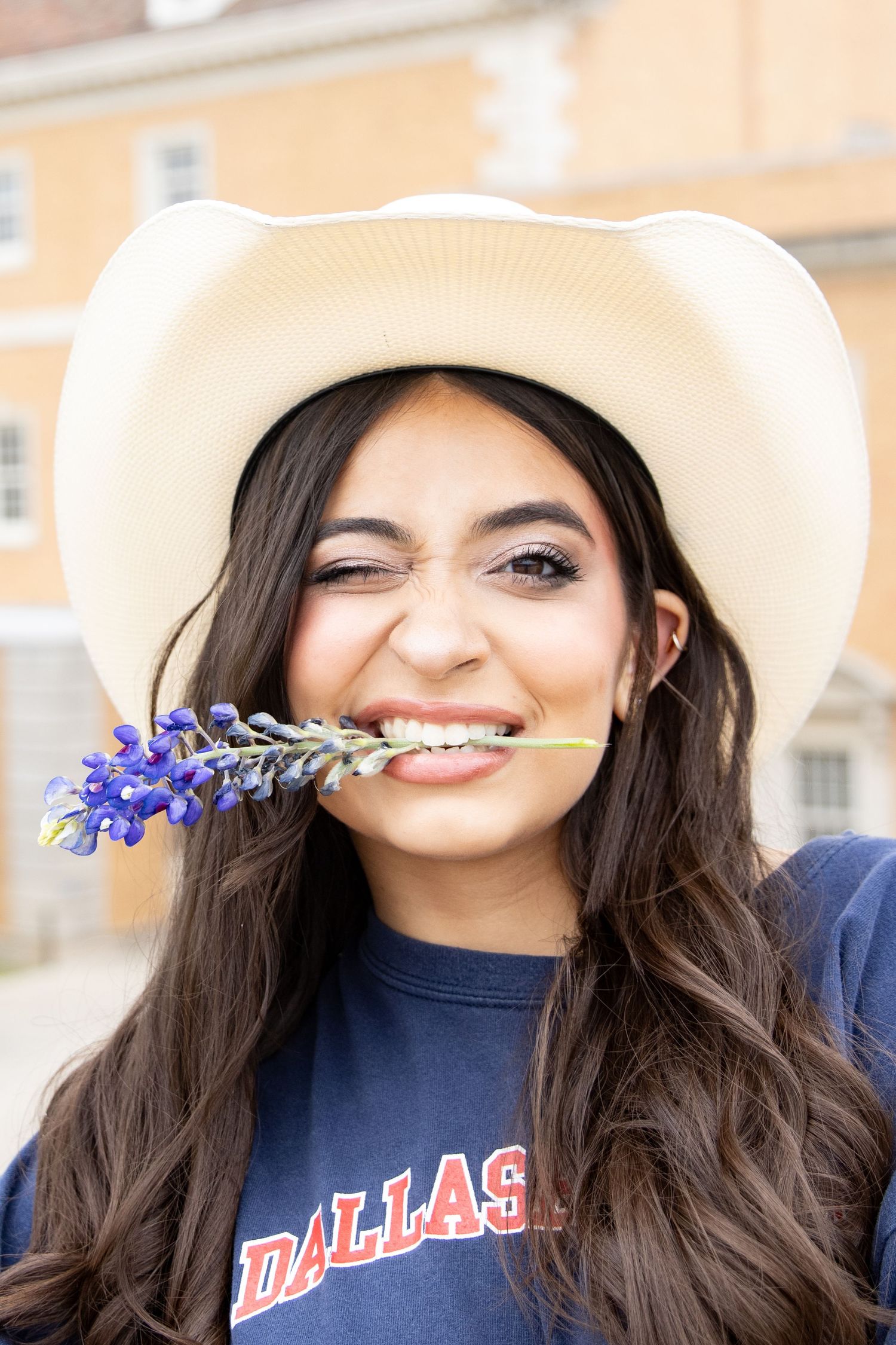 Someone wearing a Dallas t-shirt and white cowboy hat playfully holds a bluebonnet flower stem in their teeth.