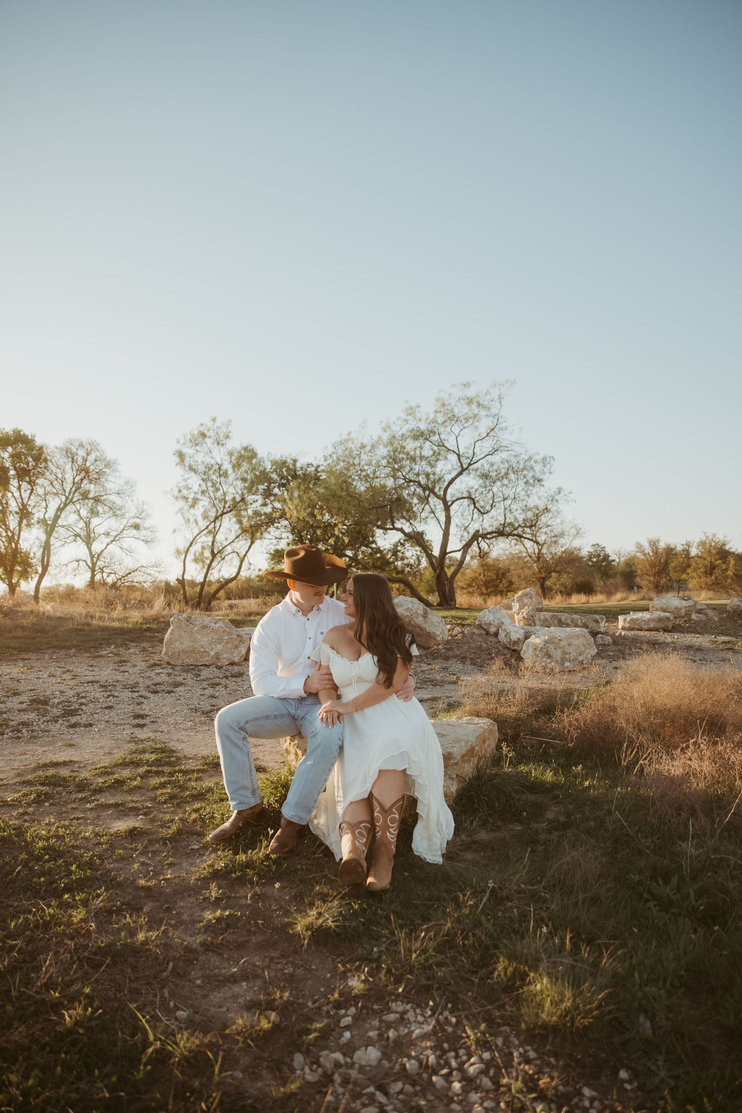 Couple in western attire sitting on a rock in a sunny outdoor setting, woman in white dress and cowboy boots.