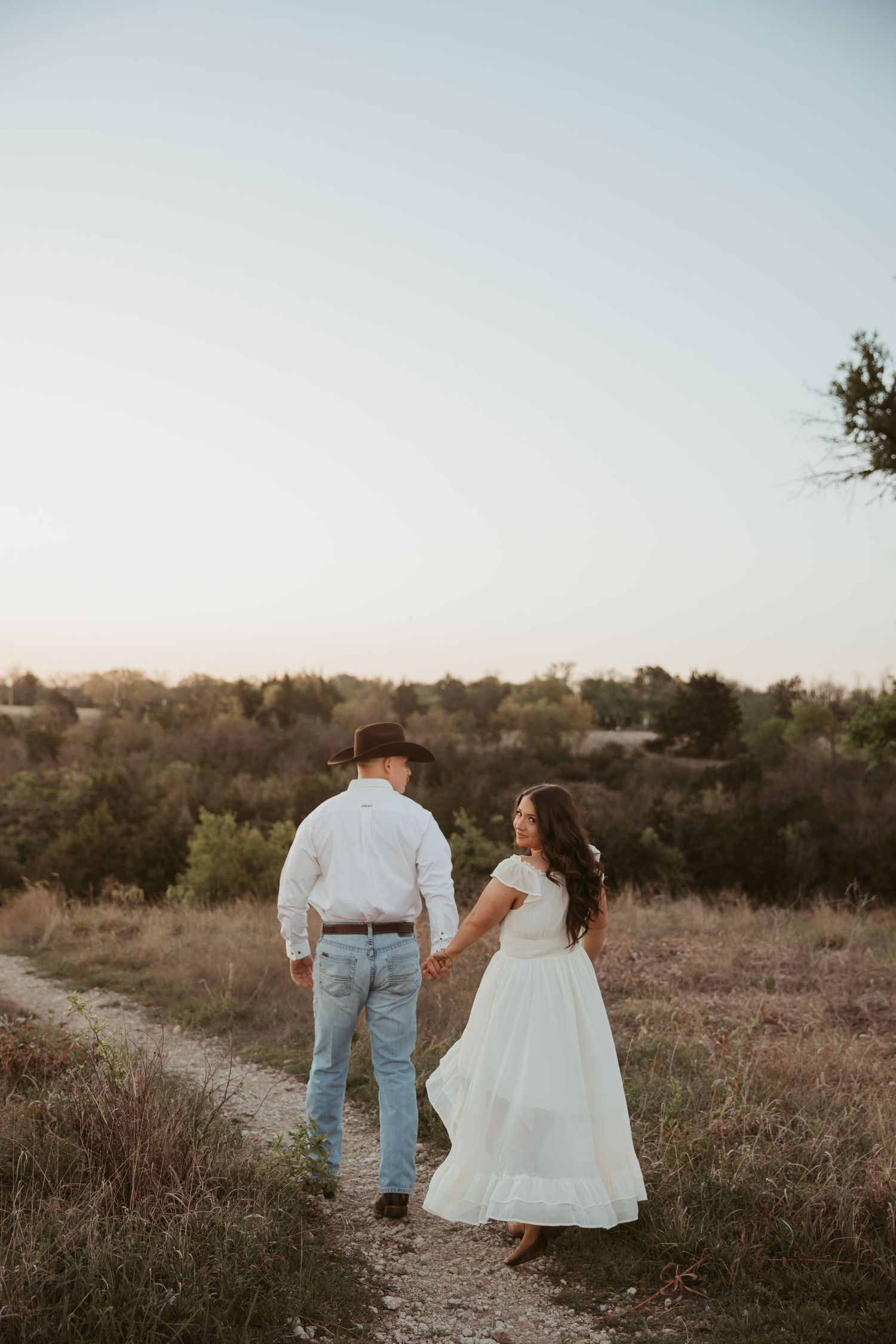 Couple holding hands walking down a rustic dirt path in golden hour light, woman in white dress and man in cowboy hat.