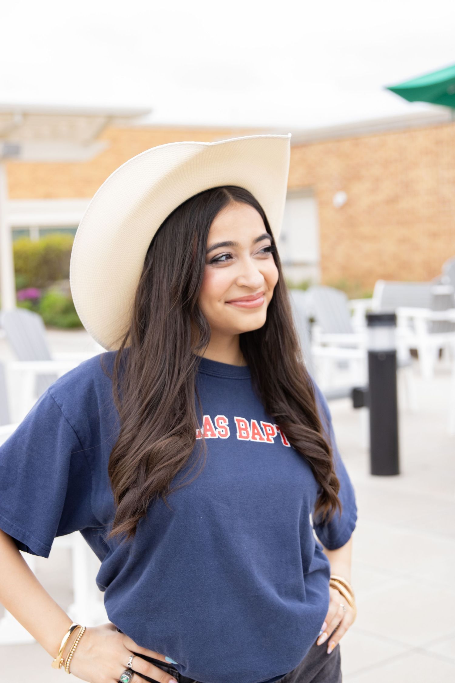 Student wearing navy t-shirt and white cowboy hat posing on campus with brick building behind.