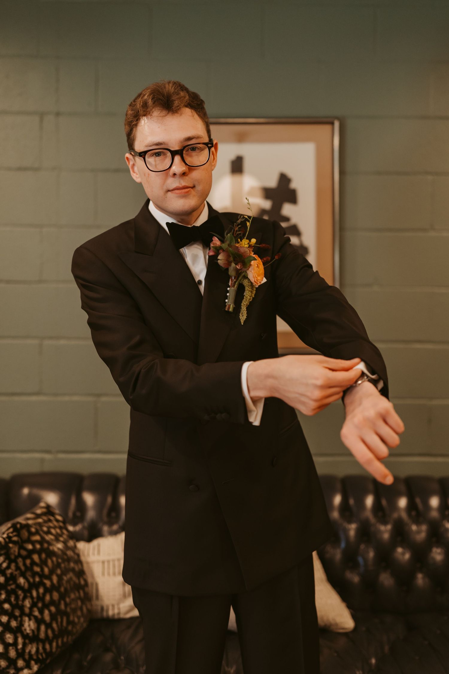 Groom in black tuxedo with floral boutonniere adjusting cufflinks before wedding ceremony.