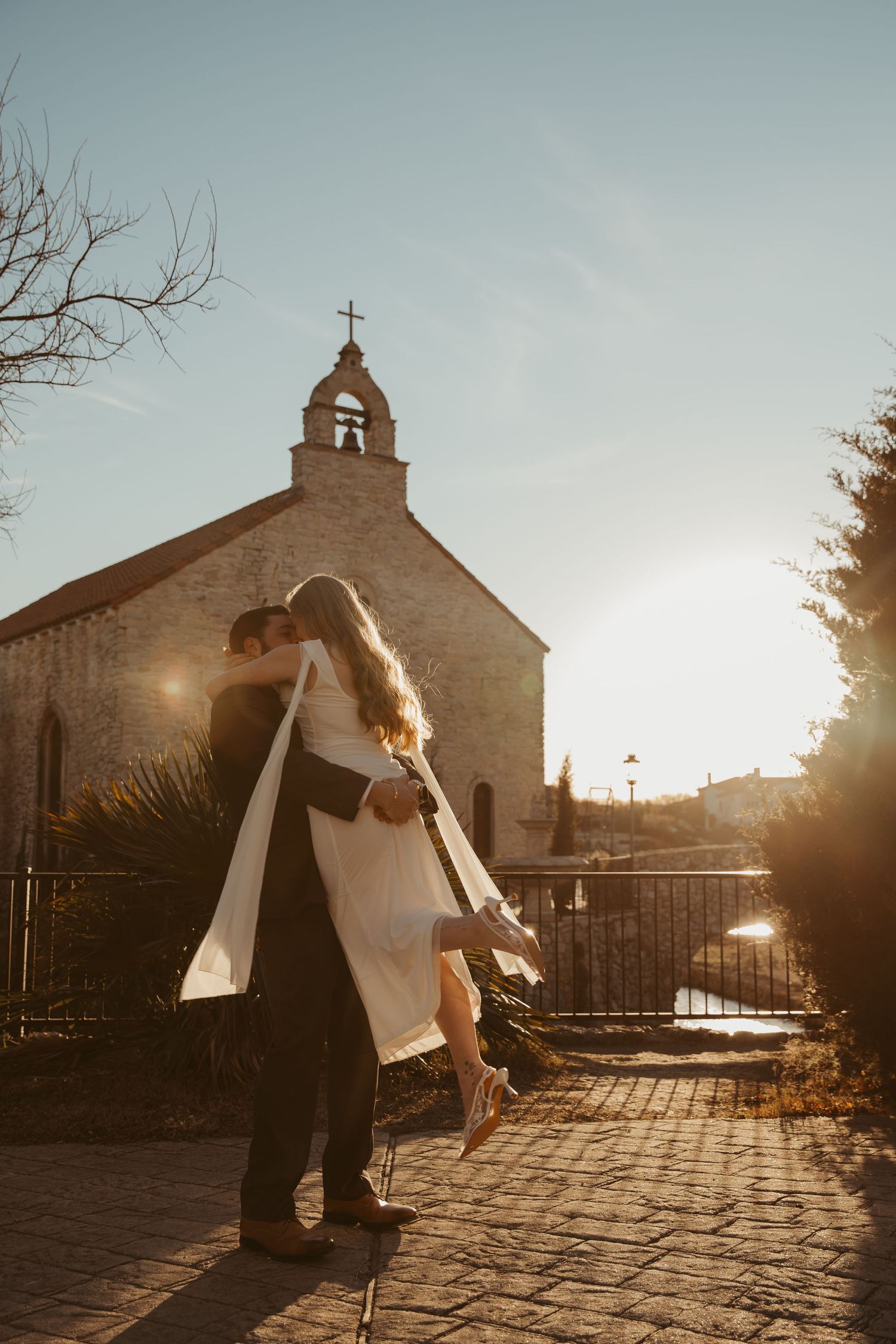 A couple shares a romantic sunset moment in front of a rustic stone church with warm golden light.