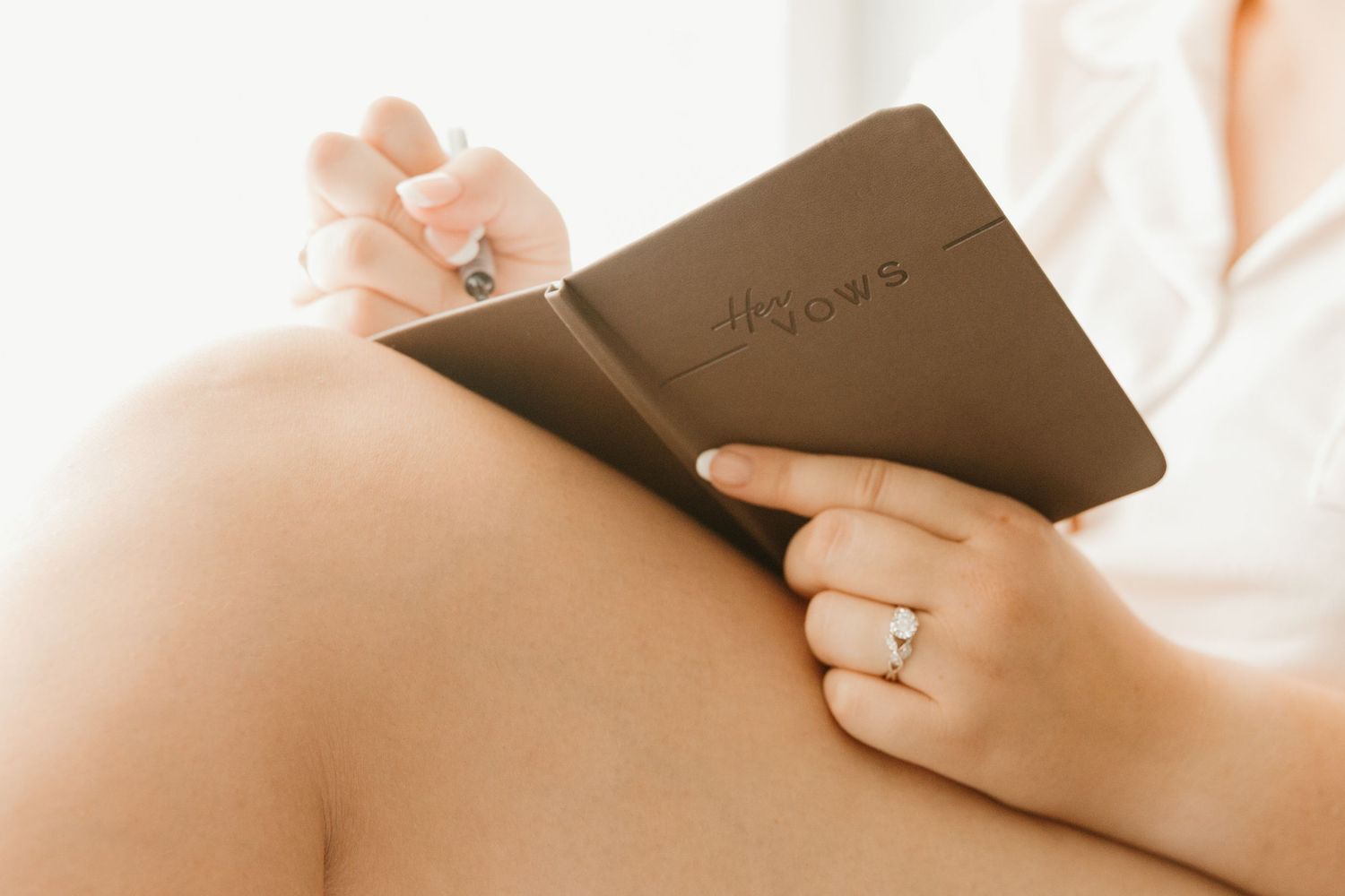 Person holding a brown leather journal while sitting on white bedding in soft natural light.