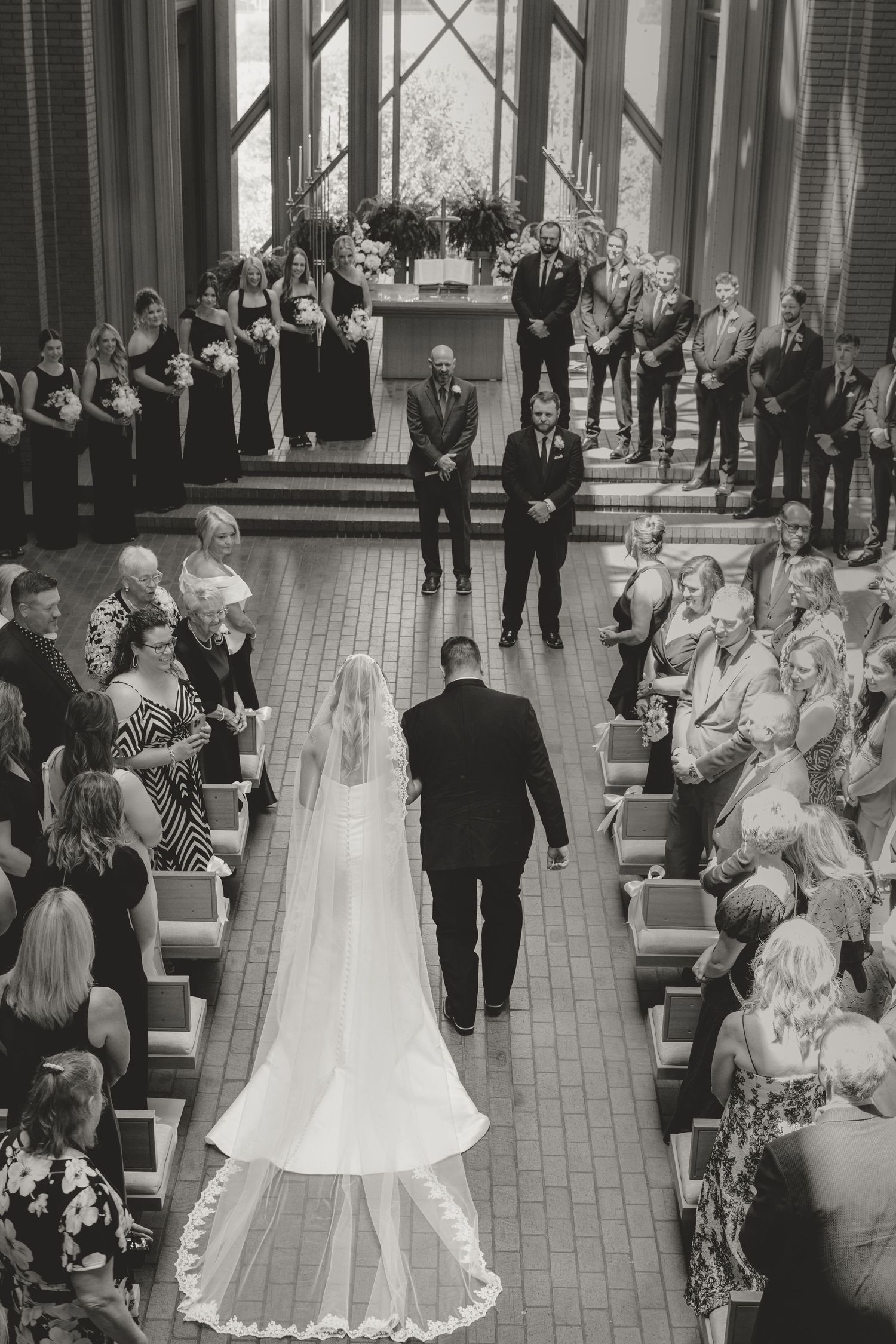 A black and white overhead view of a wedding ceremony in a chapel with guests seated in pews and the wedding party at the altar.