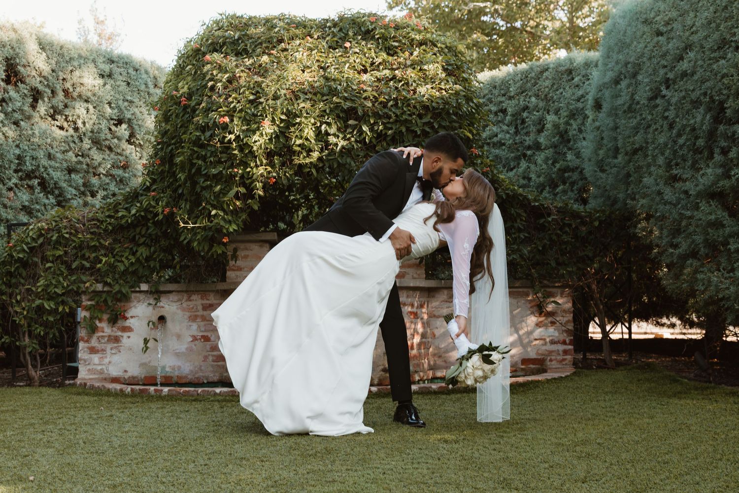 Romantic wedding scene in a garden with a couple leaning together against a vintage step ladder near green hedges.  Wedding Photographer DFW