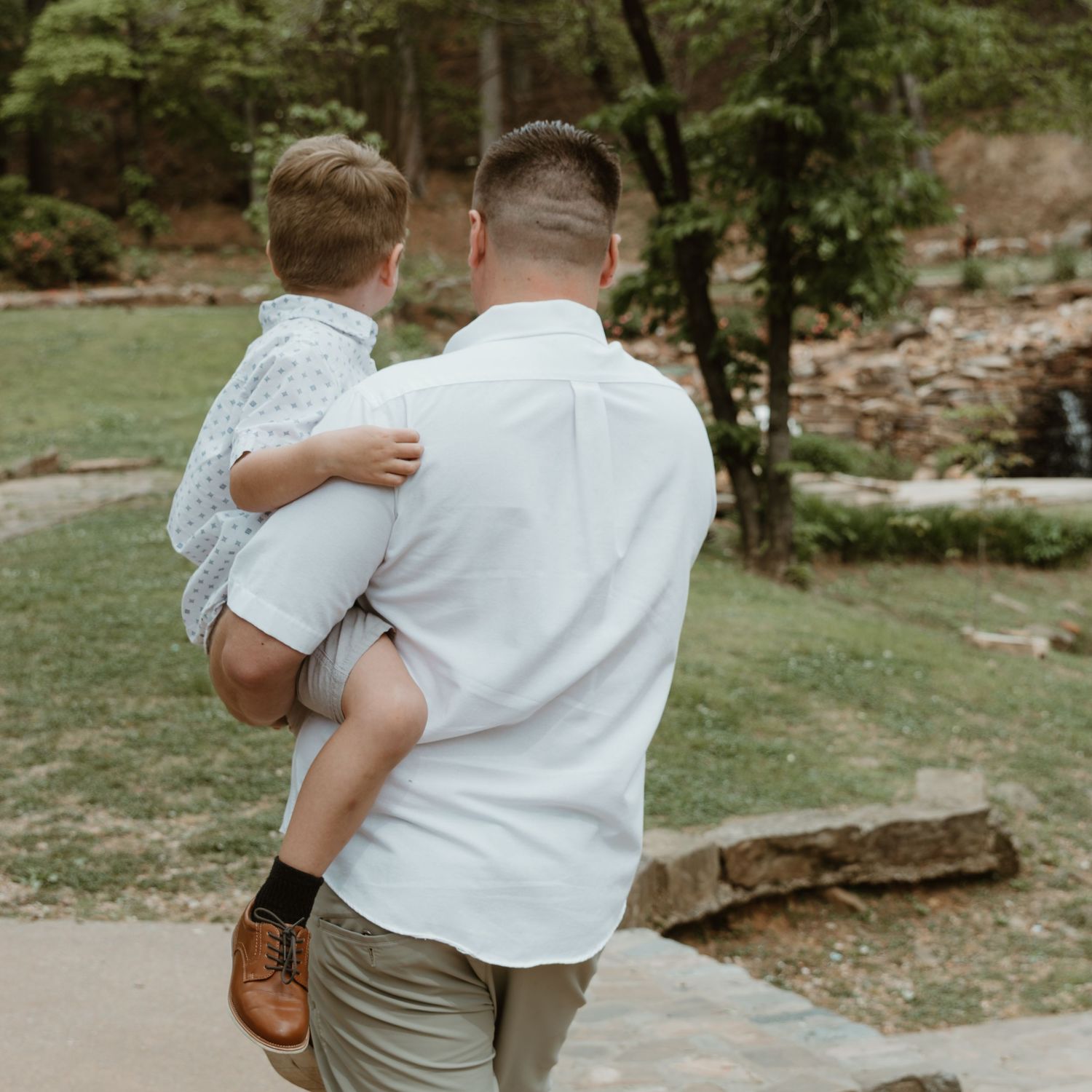 Parent in white shirt holding child while standing on stone walkway in outdoor garden setting.