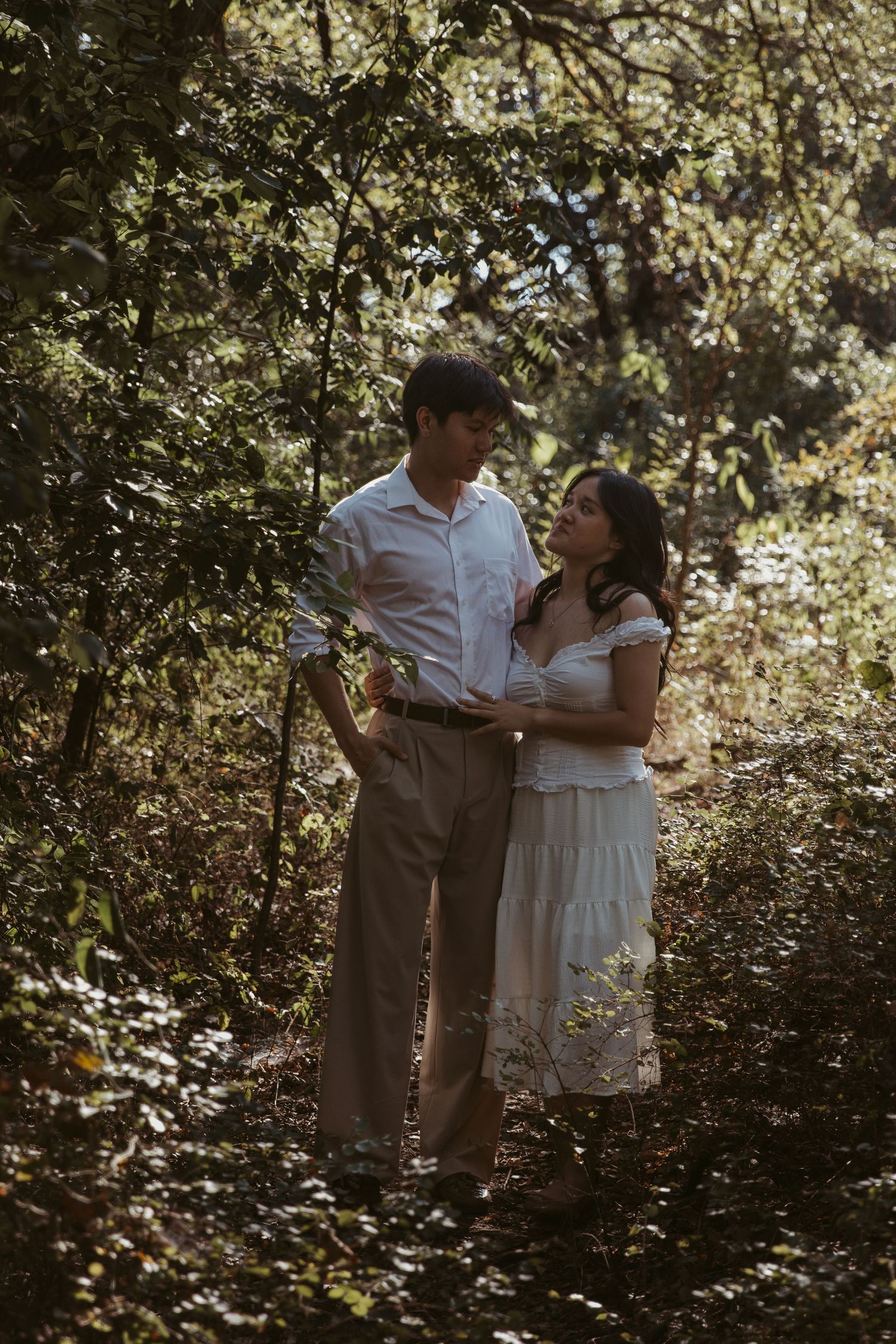 A romantic couple shares tender moments in a sun-dappled wooded area while wearing light colored outfits.