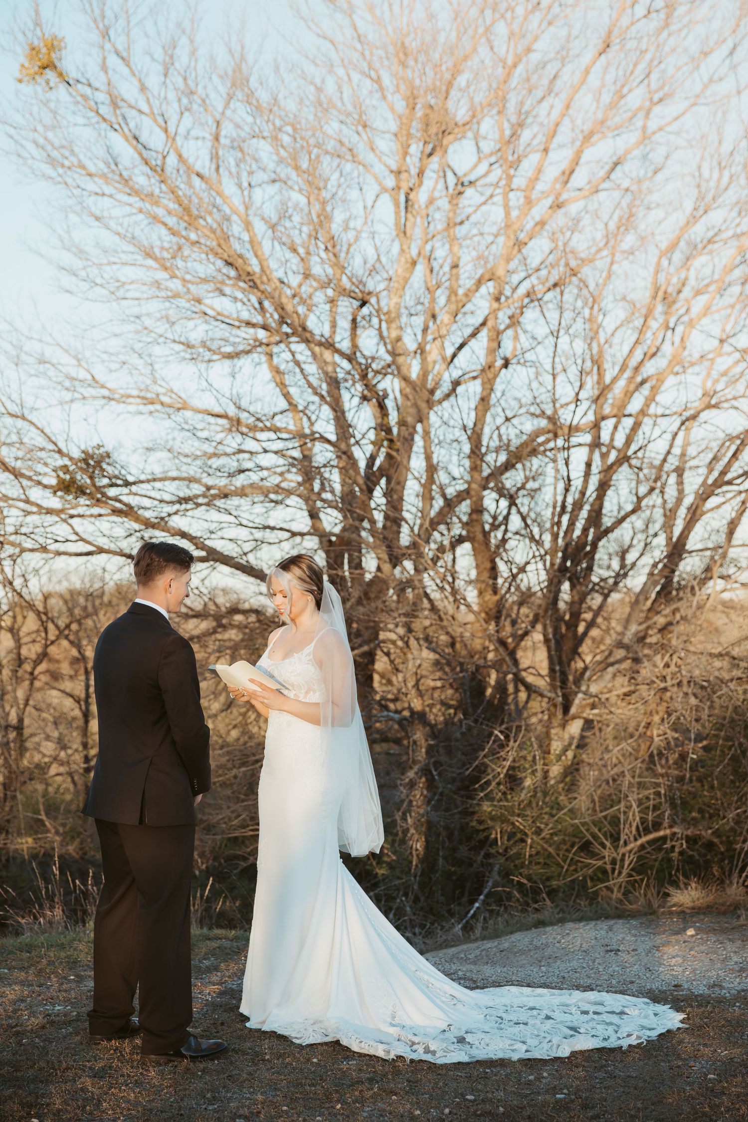 A romantic wedding couple shares a tender moment among bare winter trees at sunset.
