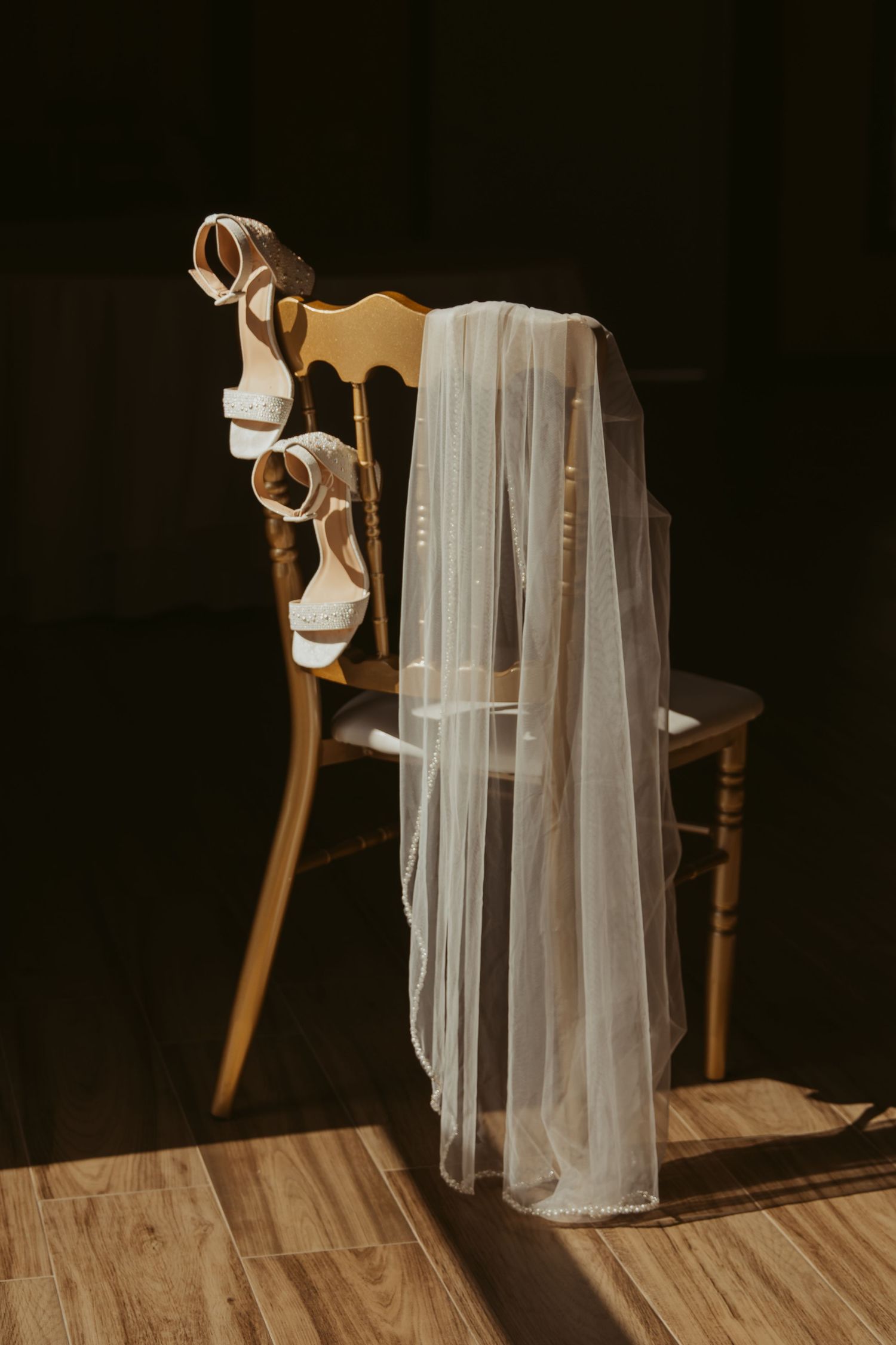 A white wedding veil draped elegantly over a wooden chair with a high heel shoe in warm natural lighting.