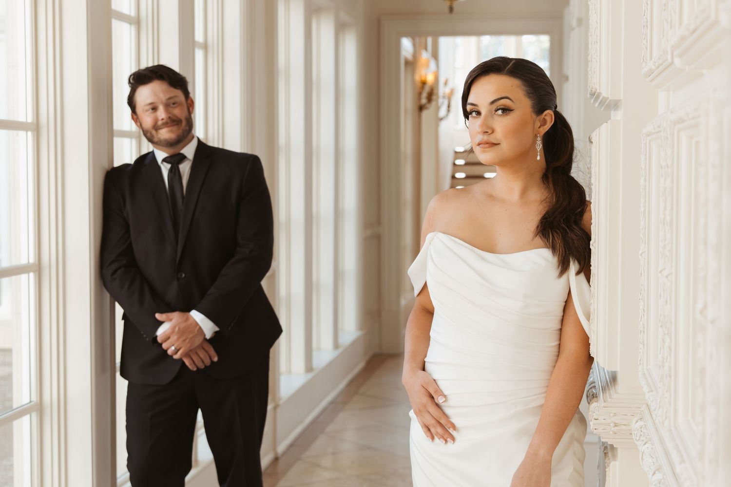 Wedding party members share a candid moment of laughter in an elegant white hallway.