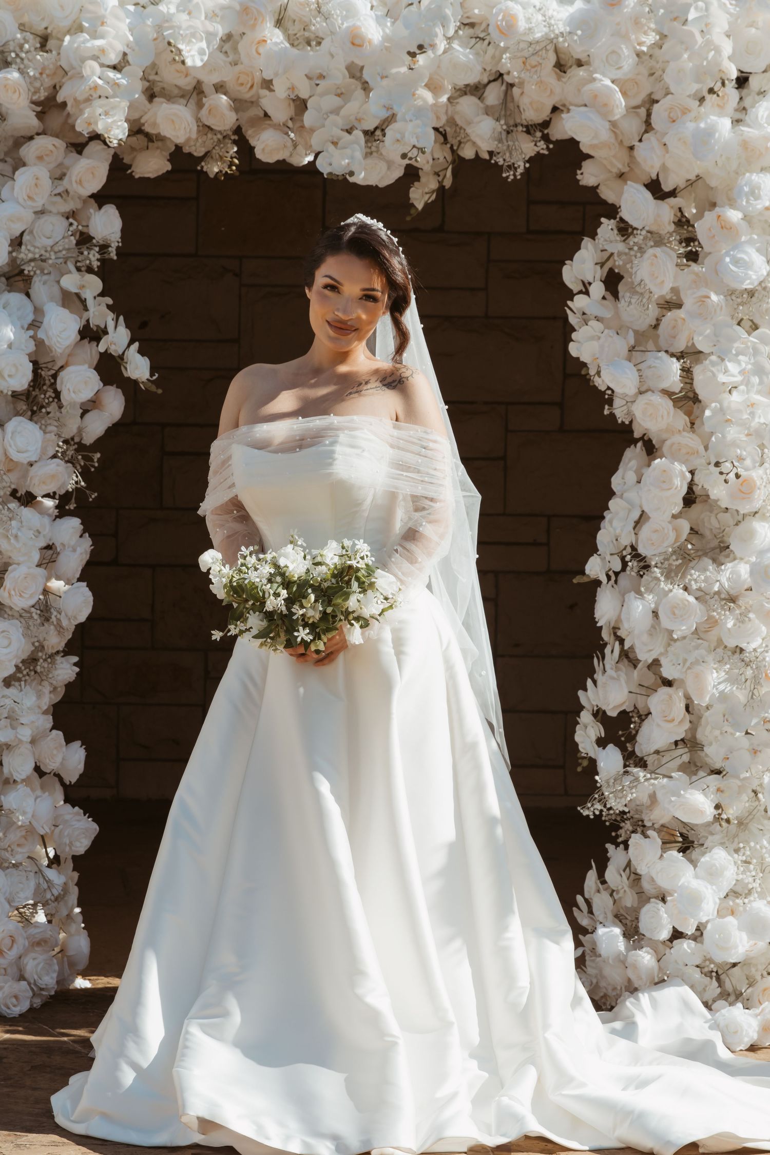 Elegant bride in off-shoulder white gown poses under floral arch with white bouquet.