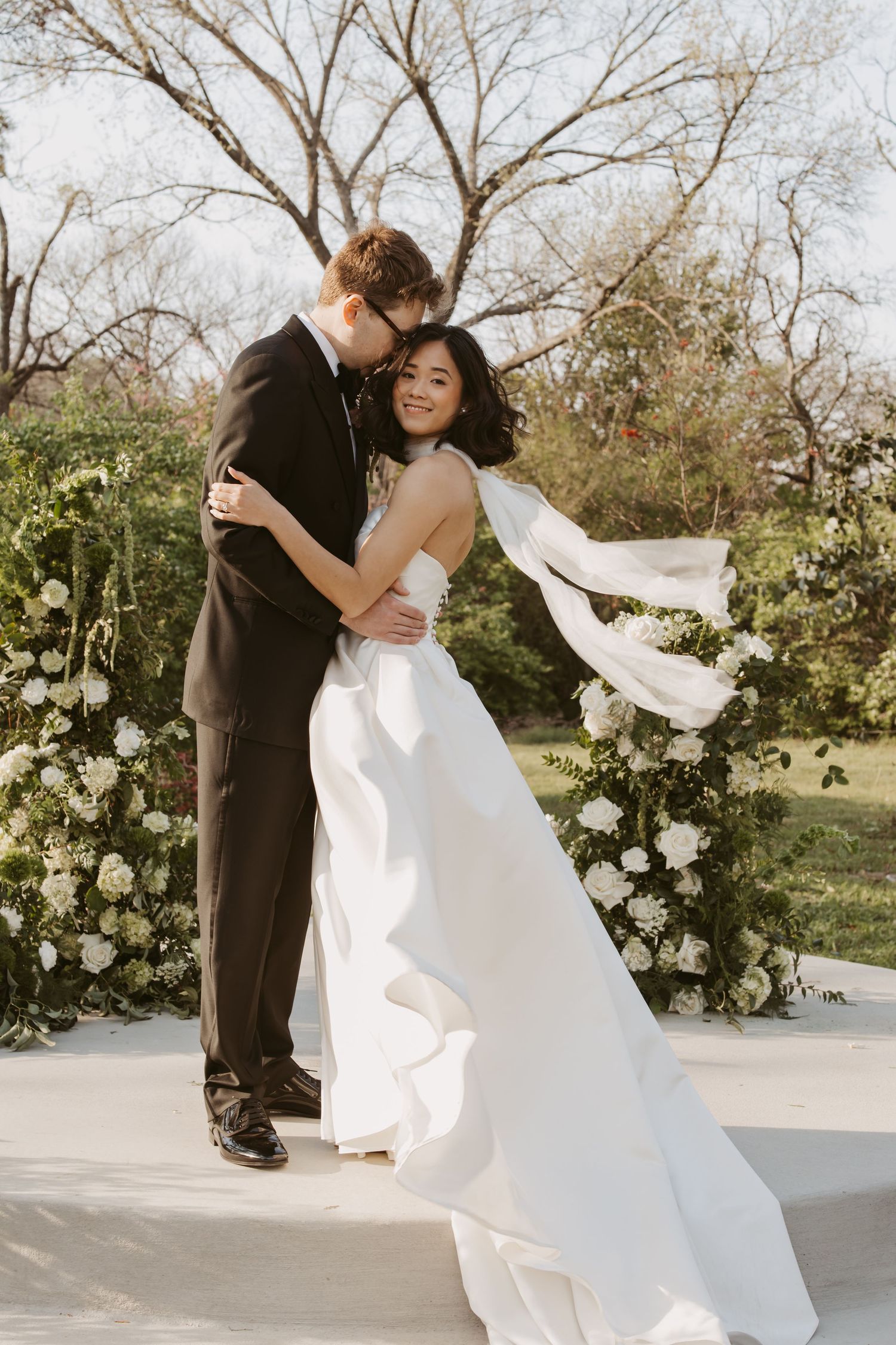 Bride in flowing white gown embraces groom in black tuxedo before a floral arch in a sunlit outdoor ceremony.