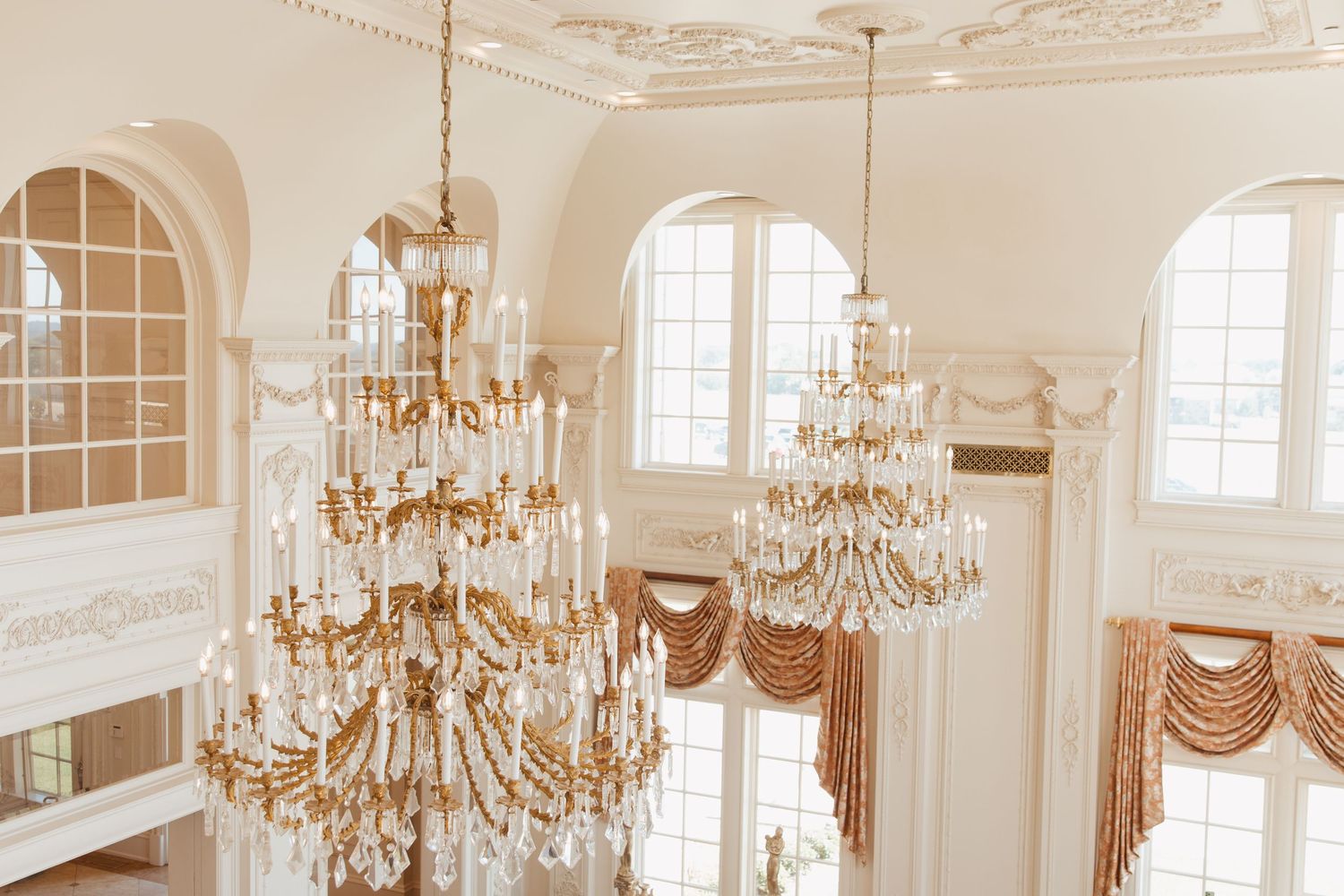 Elegant ballroom featuring ornate gold crystal chandeliers hanging from decorative white ceilings with arched windows.