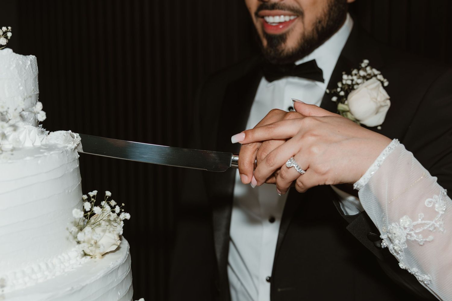 At a wedding reception, someone cuts into an elegant white wedding cake with a silver serving knife.