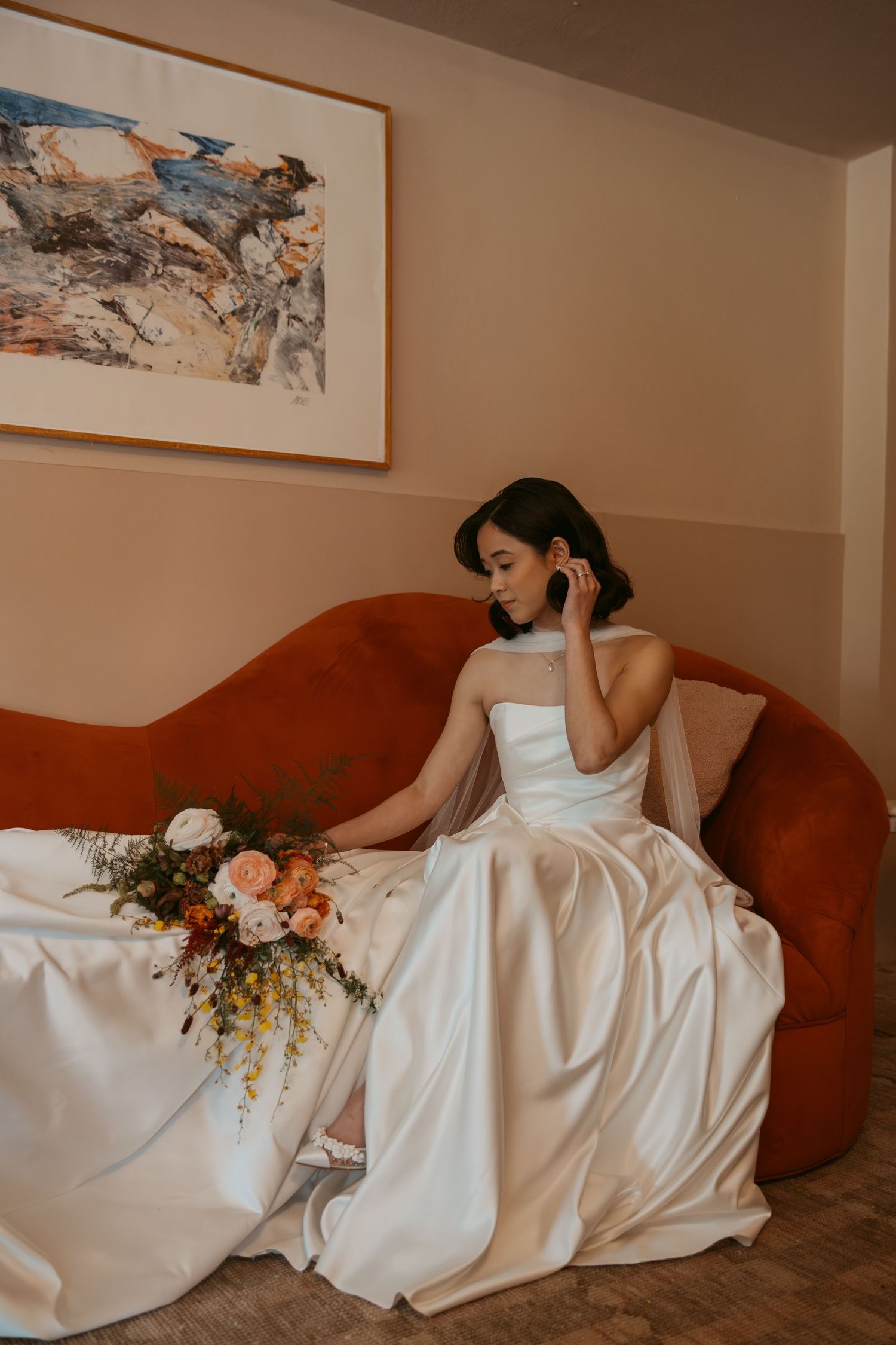 Bride in strapless white gown sits on terracotta sofa holding bouquet of colorful flowers, with abstract art on wall behind.