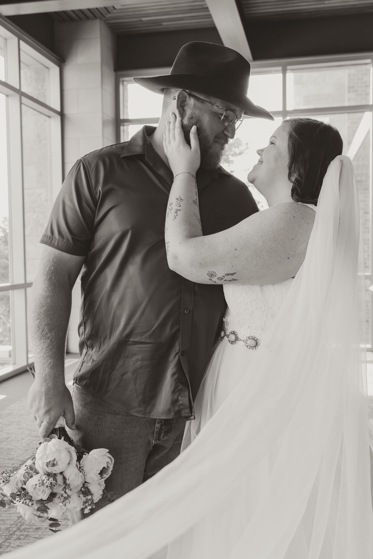 A romantic black and white wedding portrait showing a couple embracing near a window with flowing veil and bouquet.