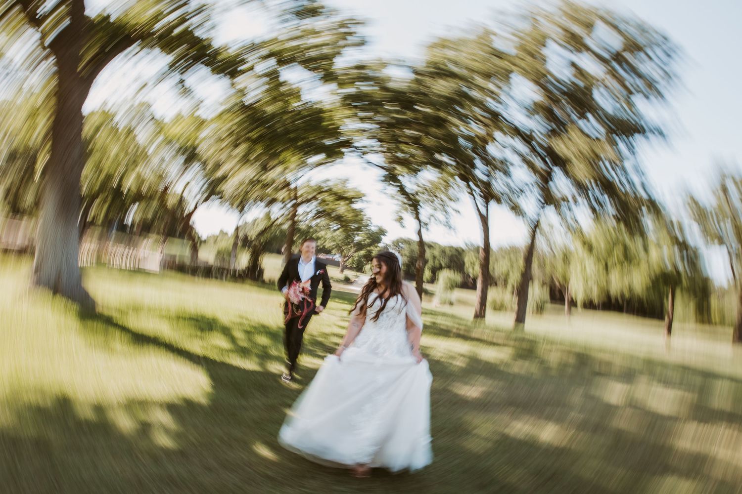 Bride and groom running through a tree-lined garden path on their wedding day, captured with a swirling lens effect.