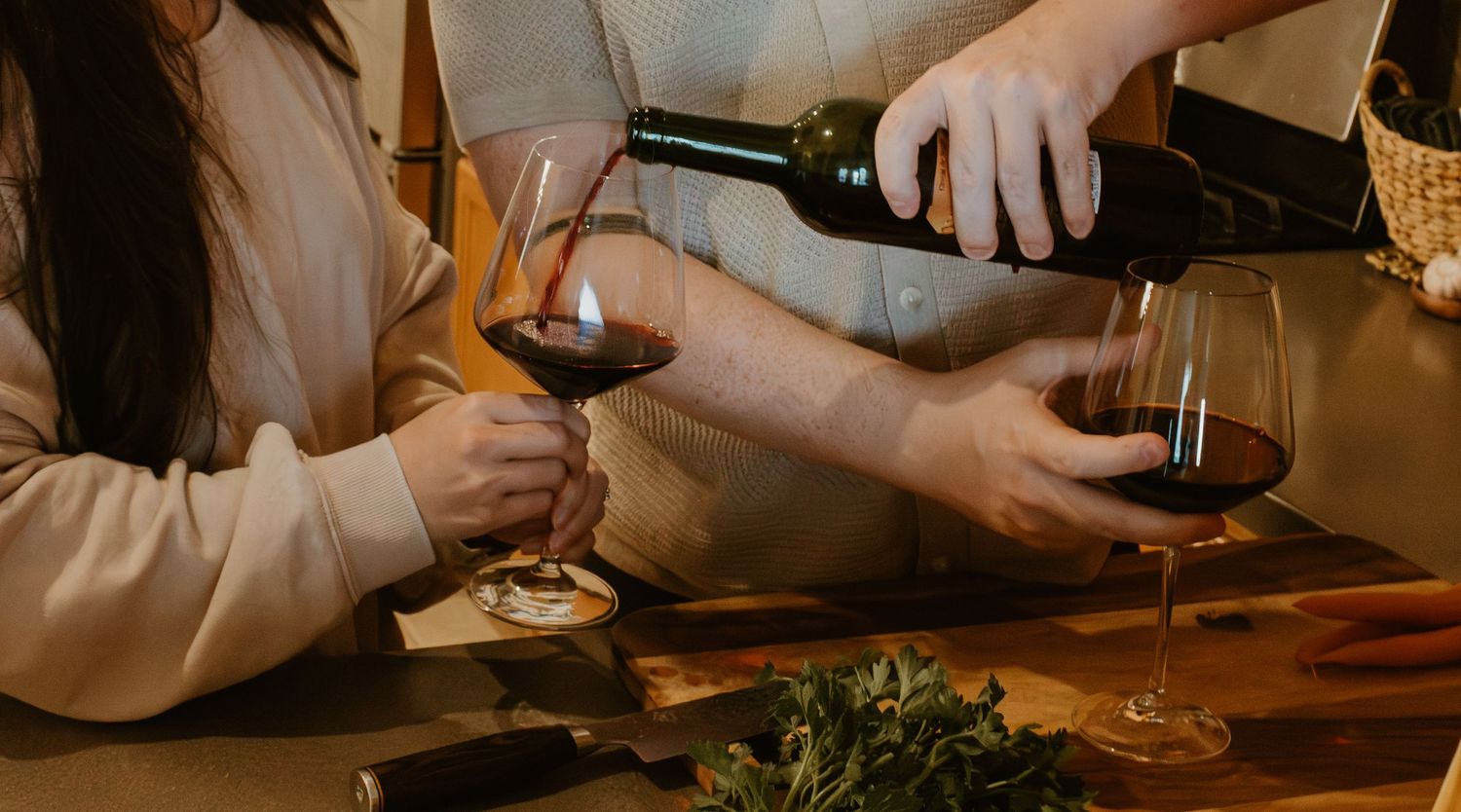 Two hands pour red wine into stemmed glasses at a wooden table setting.