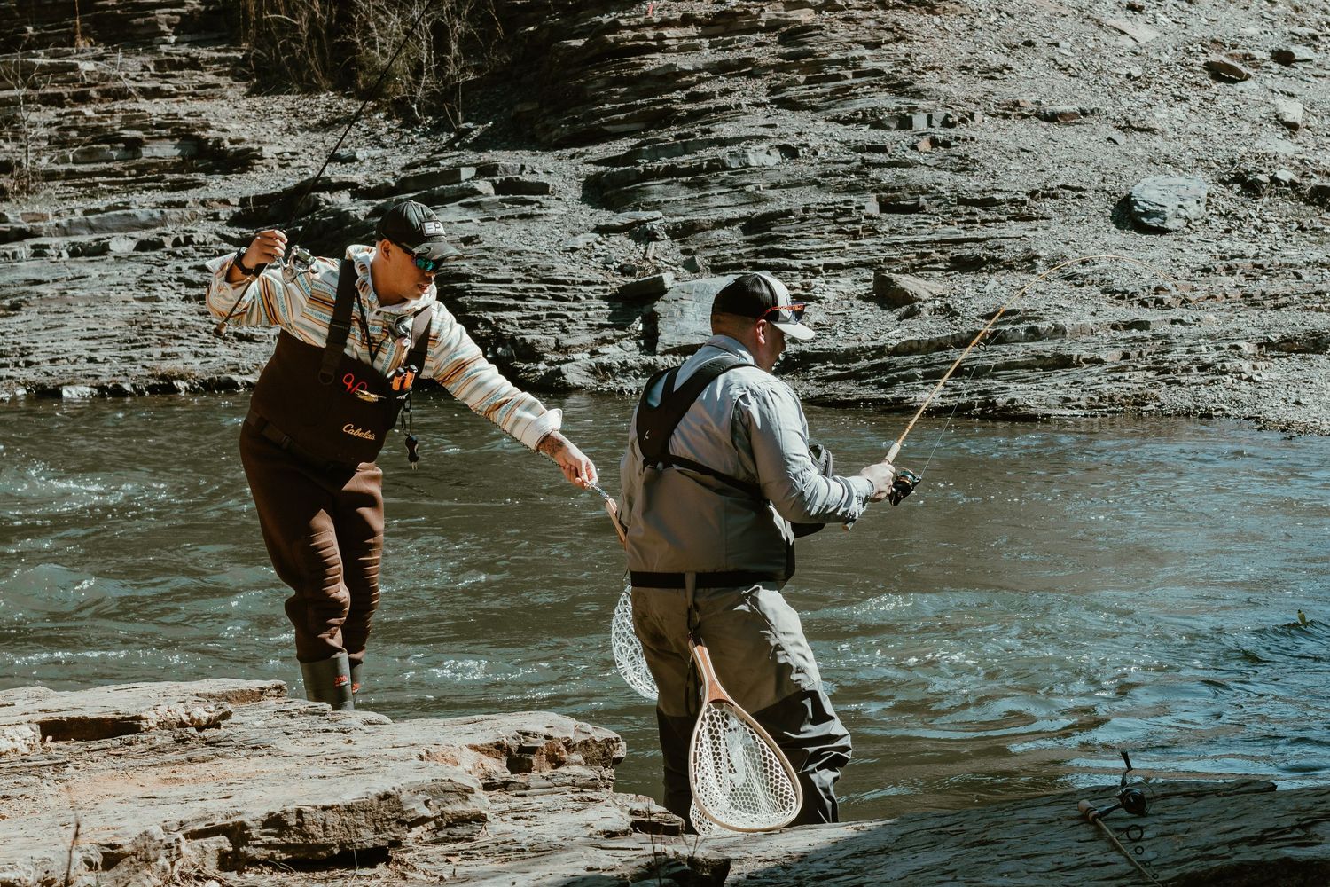 Two people in hip waders fly fishing together in a stream near a rocky cliff wall.