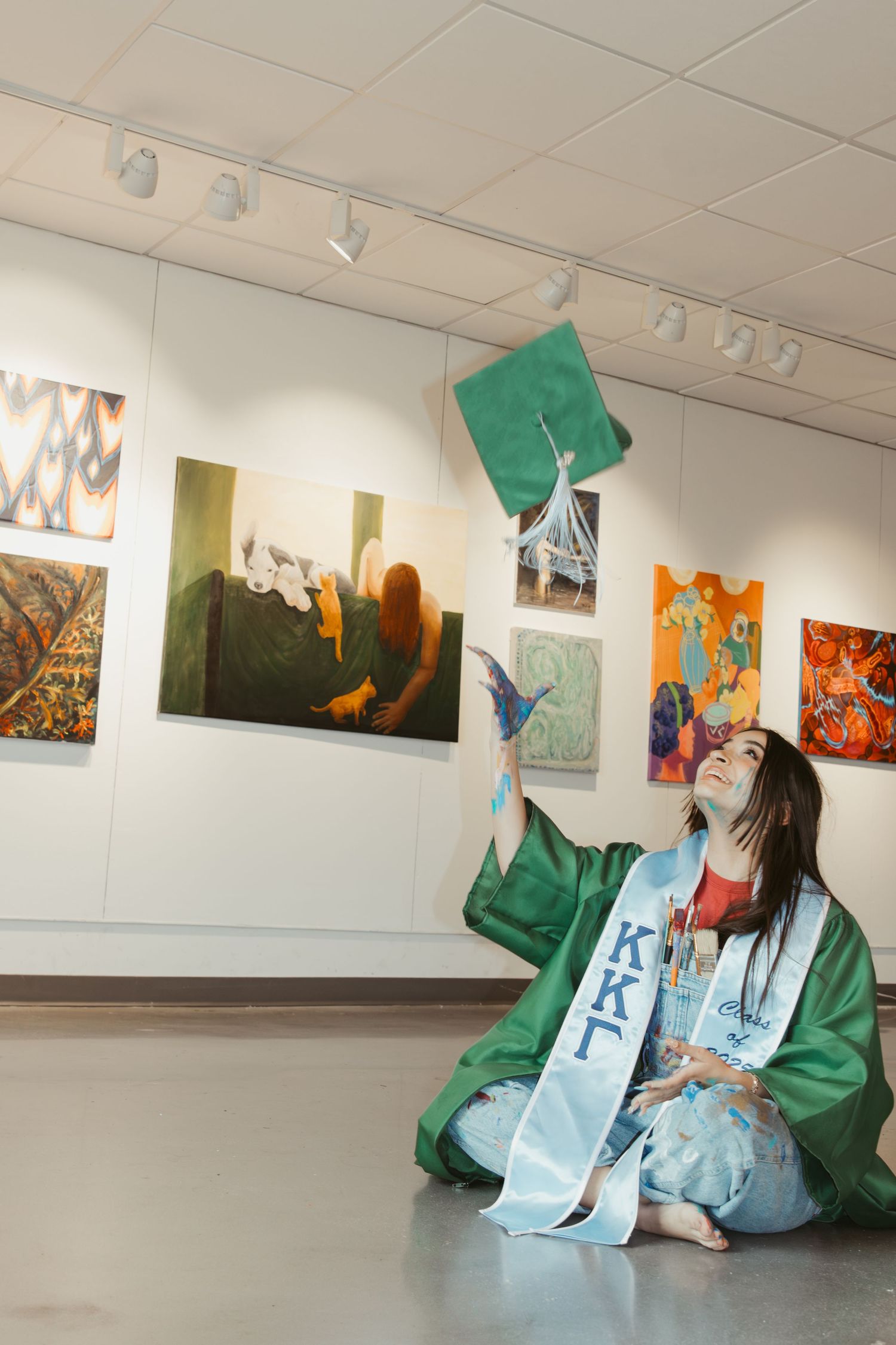 A graduate in green cap and gown sits on gallery floor surrounded by colorful artwork on white walls.