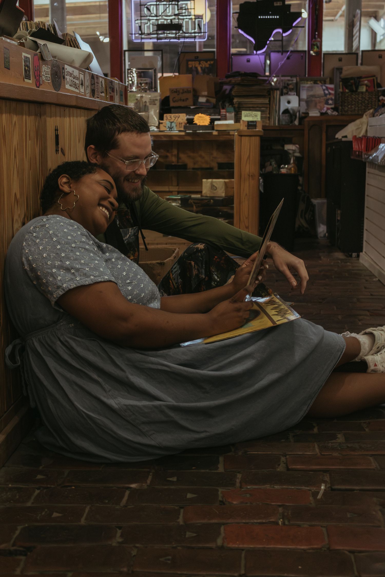 Two people sit casually on a rustic brick floor looking at something together in a cozy shop interior.