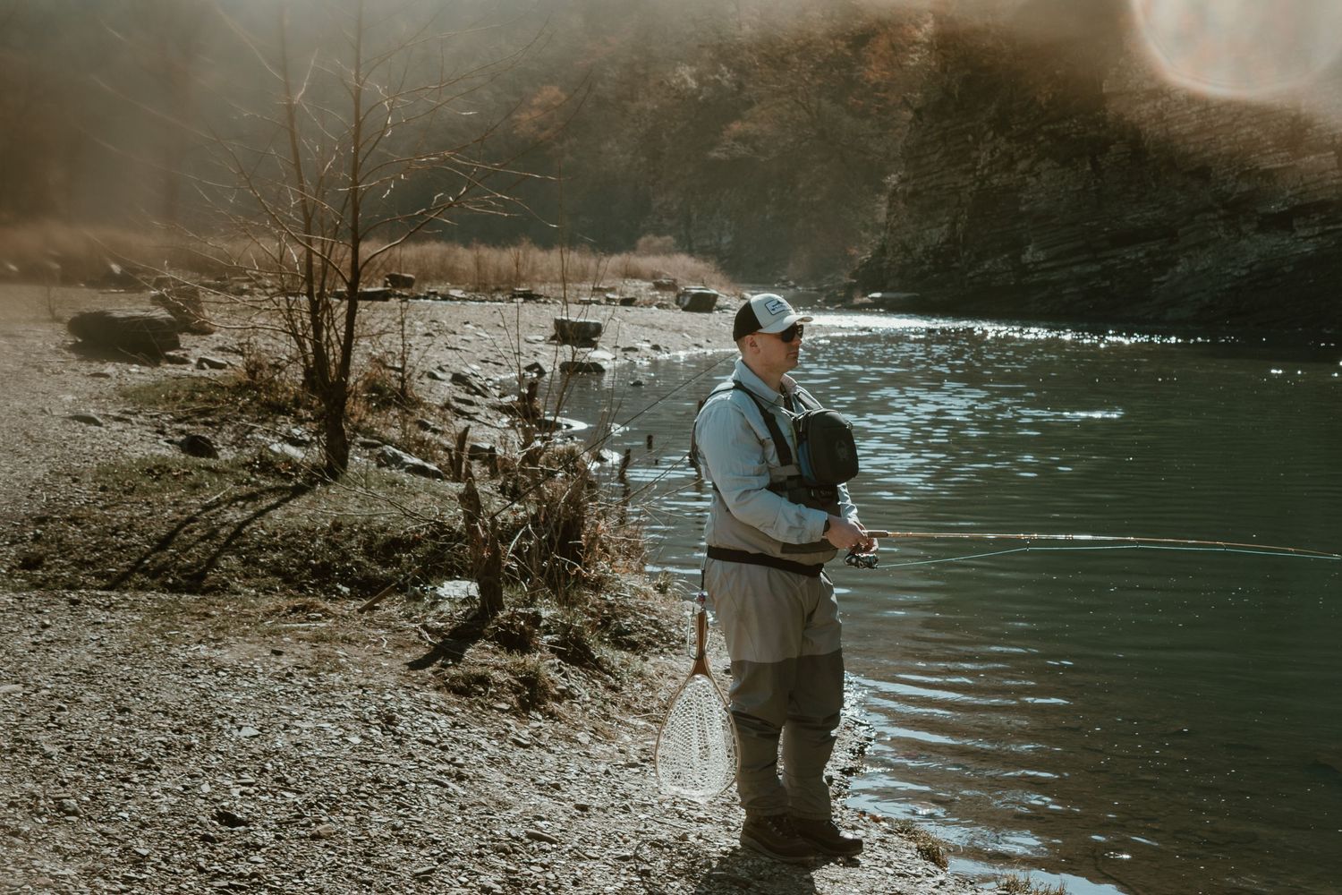 A person fly fishing stands on a rocky shoreline beside calm waters.