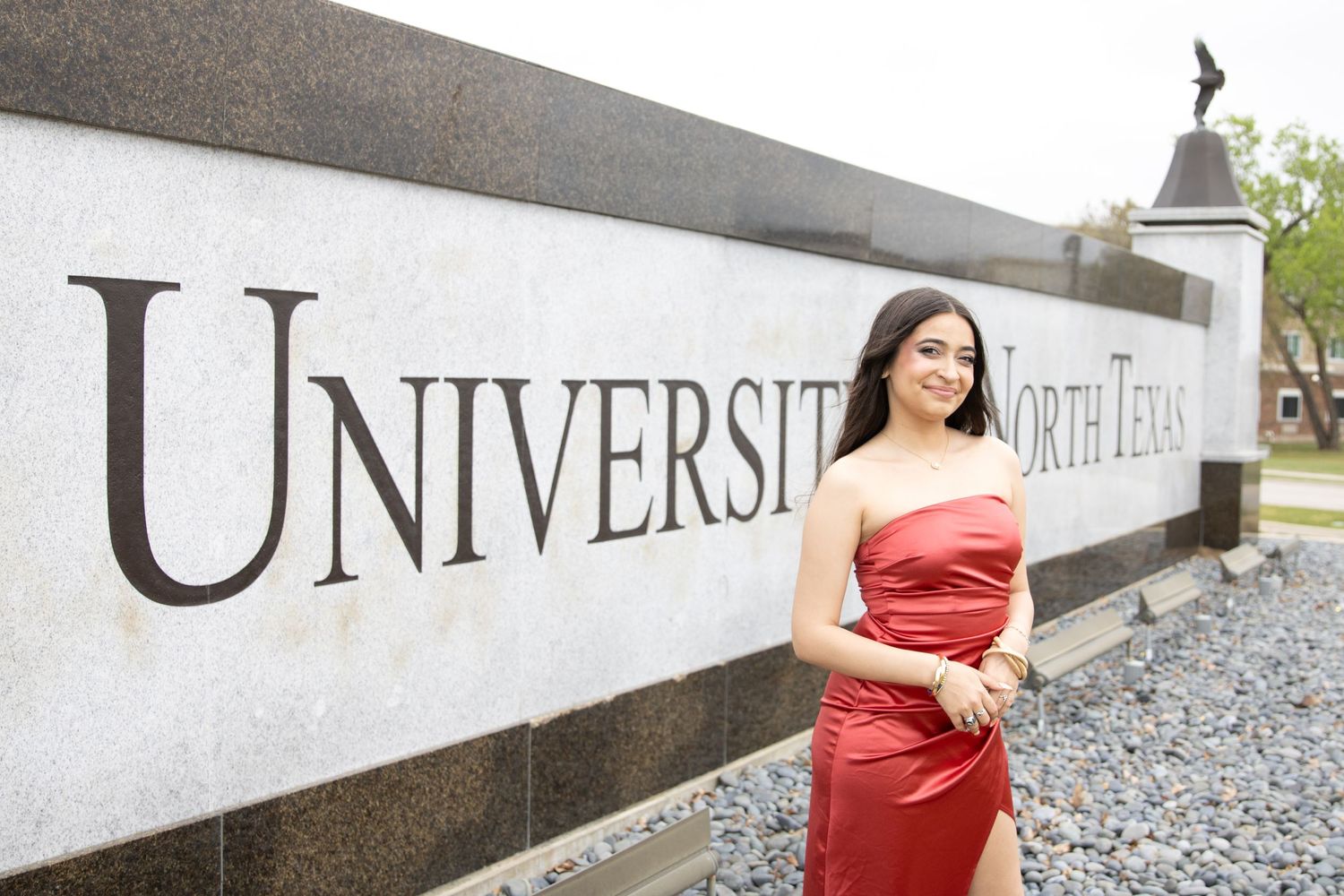 A person in a red strapless dress poses by a white university entrance sign wall.