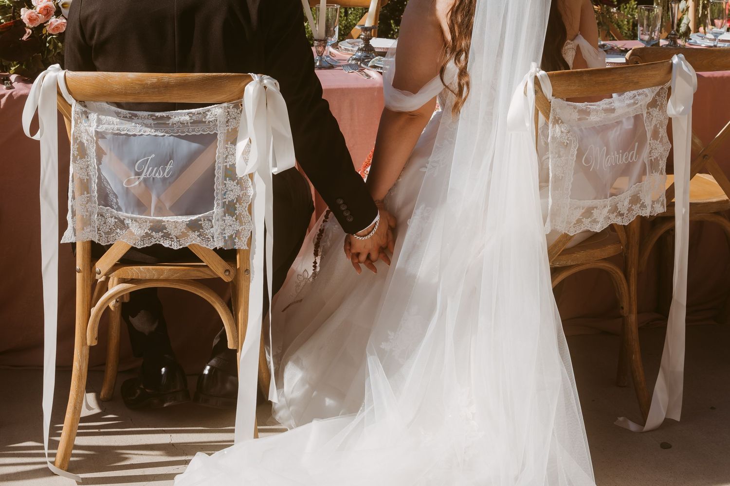 Bride and groom holding hands at wedding reception, wearing white gown with lace chair signs.