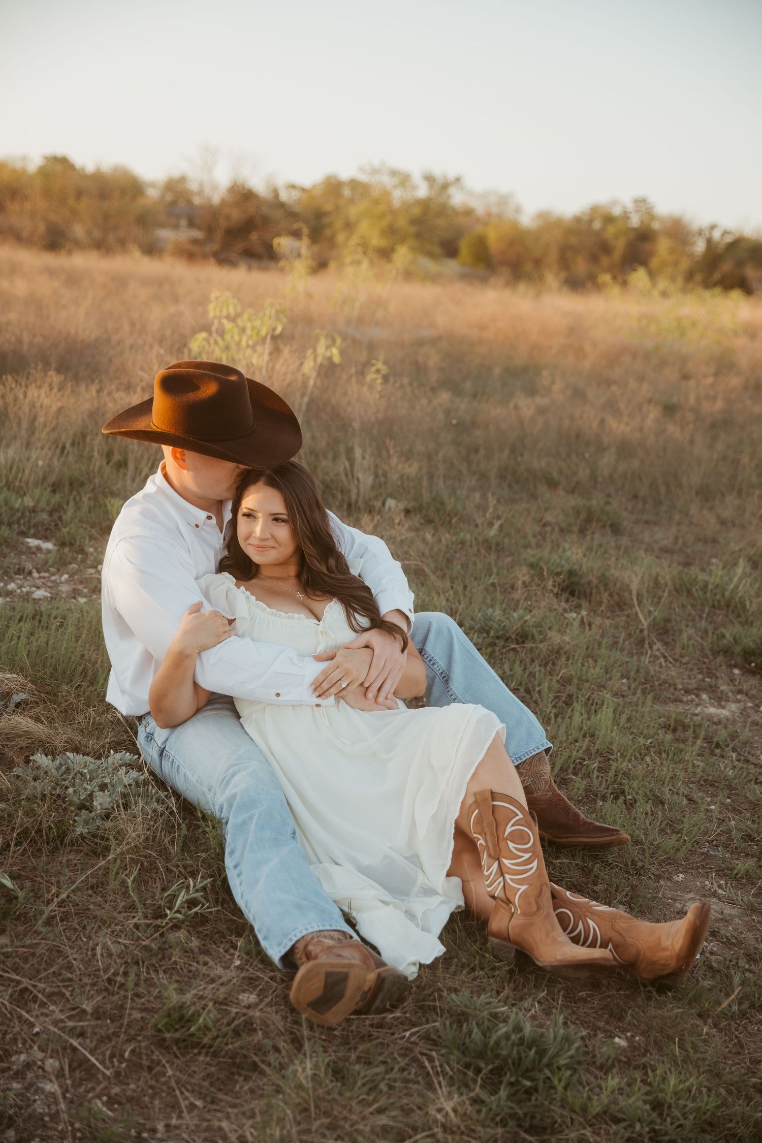Couple sitting in golden field, man in cowboy hat embracing woman in white dress and cowboy boots at sunset.